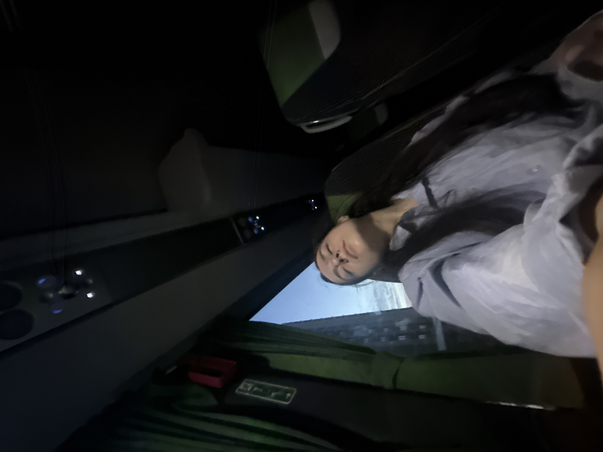 The image depicts a young woman sleeping in a dimly lit vehicle, likely a bus or train, while traveling through the city of Ostrava, Czechia. The woman, with long dark hair, is positioned in the lower center-right of the frame, eyes closed, head tilted back, and appears to be wearing a light blue, long-sleeved button-down shirt. The shot is taken from a low angle, seemingly a selfie, looking upward.
The vehicle's interior features dark upholstery on the seats, with a visible dark green upper portion of a seat to the immediate right of the woman. Overhead, a light gray panel runs across the ceiling, equipped with circular fixtures (possibly speakers or vents) and a series of small, illuminated lights, some glowing blue and others white, characteristic of public transport cabins. Green curtains partially cover a window on the left side of the frame, with a red strap or handle hanging near them. Through the window, an urban building with multiple dark rectangular windows is visible against a light blue sky with faint cloud patterns, suggesting either dawn or dusk. The overall lighting within the vehicle is subdued, contrasting with the soft natural light visible outside. No direct interactions between individuals are depicted, and the primary action is the woman resting.