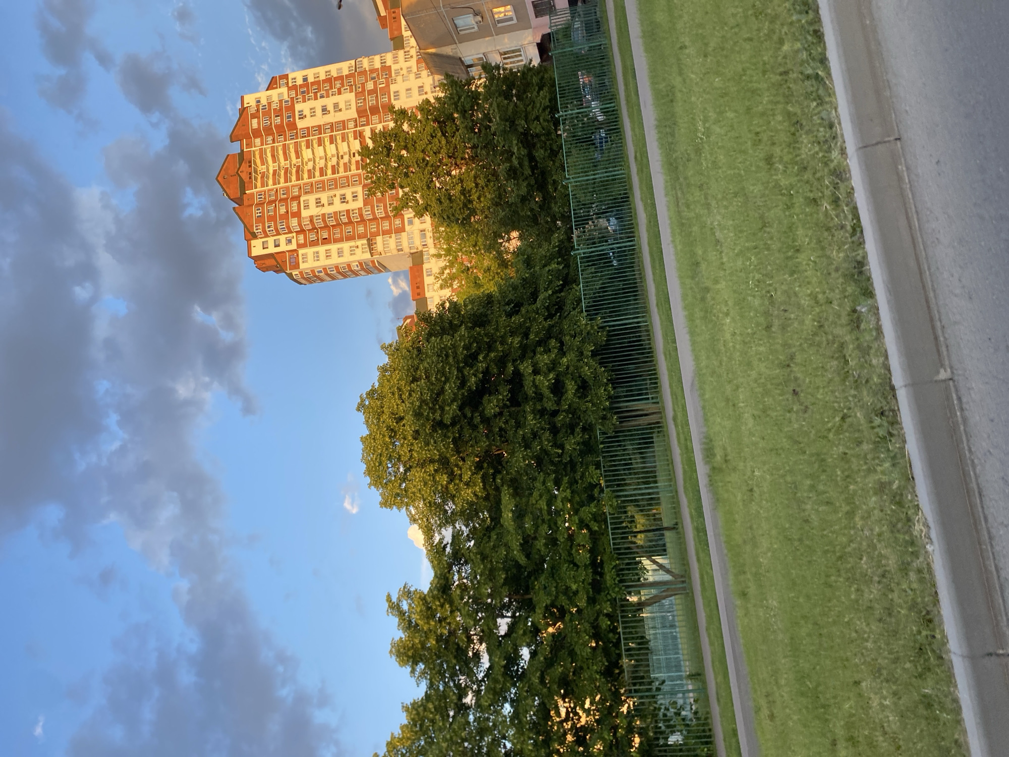 The image captures an outdoor scene featuring a prominent residential building and natural elements under a partly cloudy sky. The primary subject is a tall, multi-story apartment building situated on the left, characterized by its reddish-brown and white facade, numerous windows, and a distinctive, stepped roofline. The building's side facing the viewer is illuminated by warm, golden sunlight.
In front of and to the right of the building, a dense cluster of large, leafy green trees occupies the mid-ground, also catching the sunlight, which suggests a late afternoon or early evening time of day. Above, the sky is a blend of clear blue and scattered, soft grey clouds.
To the right of the trees and extending into the foreground is a clear landscape section. This includes a wide strip of vibrant green grass, bordered on its left by a green metal fence and on its right by a paved sidewalk and a dark grey road with a concrete curb. Another, smaller building with a greyish-pink facade is partially visible behind the trees and further back on the right. No people or specific activities are visible in the scene. There is no discernible text. The setting appears to be an urban or suburban area.