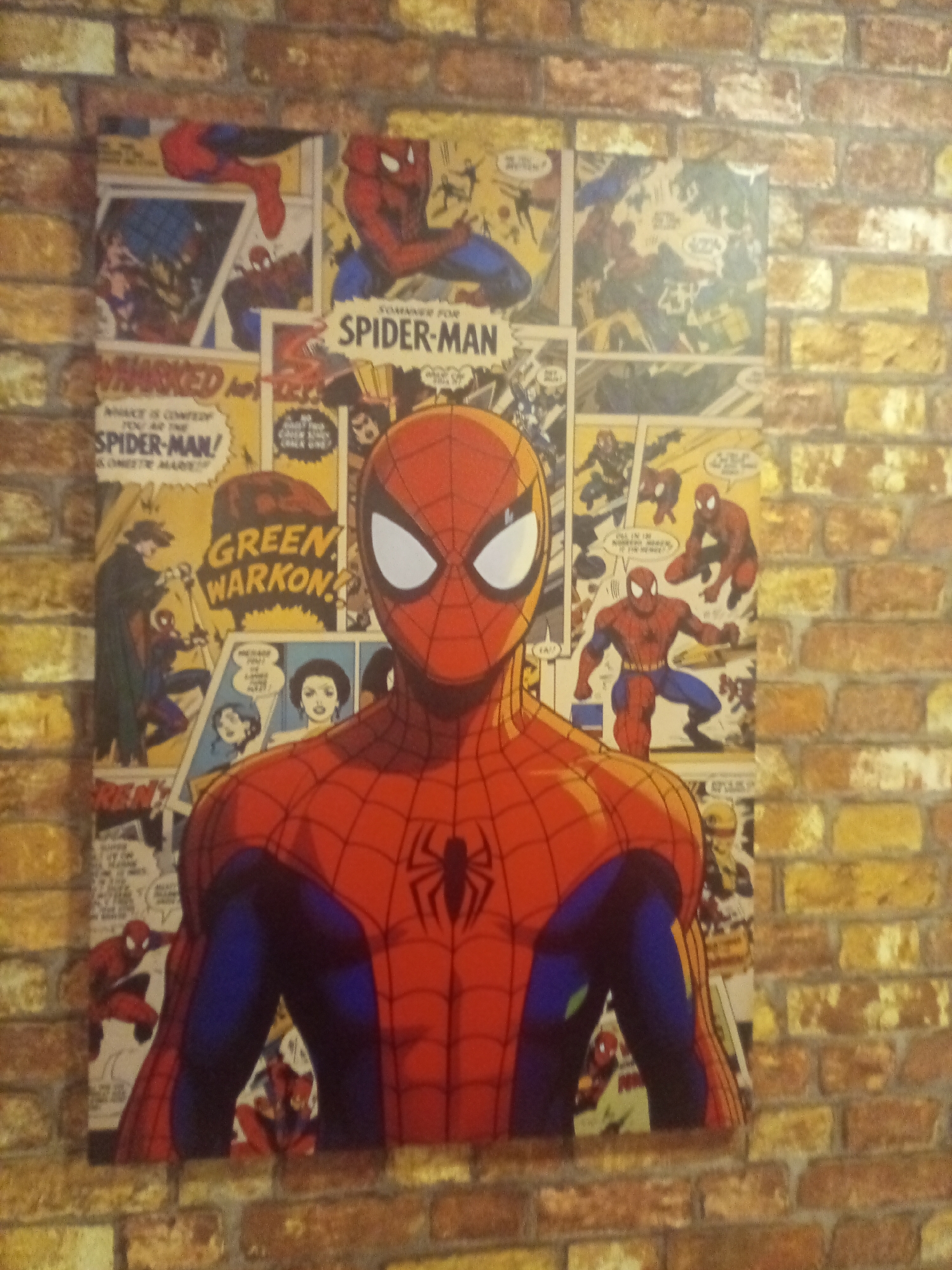 A vertical, close-up shot captures a Spiderman comic book mural displayed on a brick wall. The mural features a large, prominent image of Spiderman in the foreground, his iconic red and blue suit detailed with its web pattern. Behind Spiderman, multiple panels from various comic books are arranged, showcasing different scenes and depictions of the superhero in action, as well as other characters like Green Goblin. The brick wall, with its varied shades of orange, brown, and yellow bricks, provides a textured backdrop to the vibrant mural. The lighting appears to be ambient, casting a soft glow on the scene. The mural is likely located indoors, given the texture of the wall and the way the light falls. The specific location in Porlamar, Venezuela, is not determinable from the image.