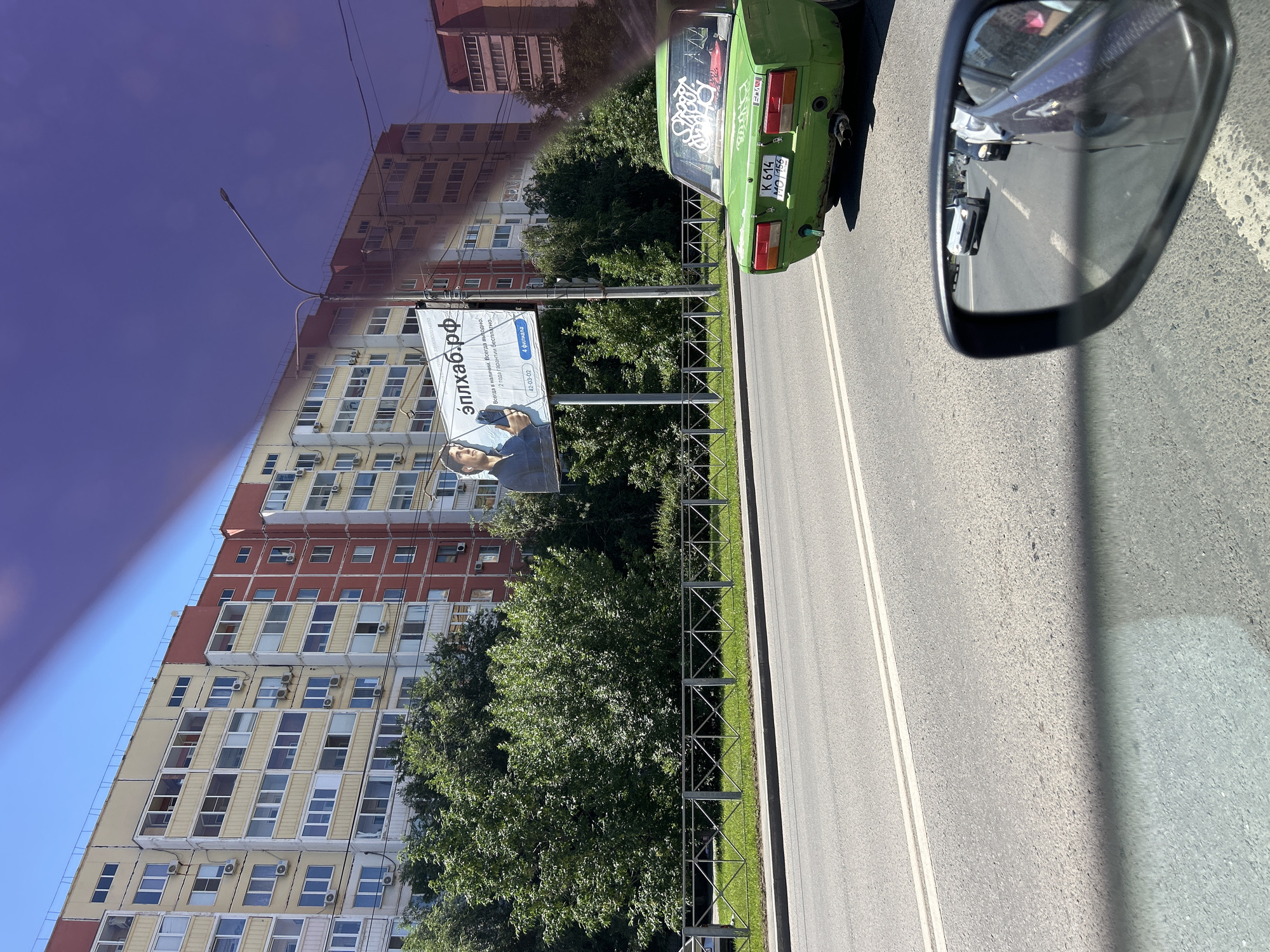 The image shows a street scene in Orenburg, Russia, likely taken from inside a vehicle. A green car with graffiti on its rear window is parked on the right side of the road. In the background, a tall apartment building with many windows and balconies dominates the scene, with trees in front of it. A billboard is visible, displaying text and an image of a person.
The text on the billboard is in Russian and reads "эплхаб.рф" (eplhab.rf), which is likely a website address. Below this, it says "Всегда в наличии. Всегда выходно" (Always in stock. Always convenient) and "2 года гарантии бесплатно." (2 years of free warranty.). There are also phone numbers and the word "филиала" (branches). The license plate of the green car reads "K 614 MO 156".
The scene appears to be set during the daytime, with bright sunlight casting shadows. The sky is a deep blue, suggesting clear weather. The overall impression is of an ordinary urban environment in Russia. The image is captured at an angle, possibly indicating it was taken while moving.