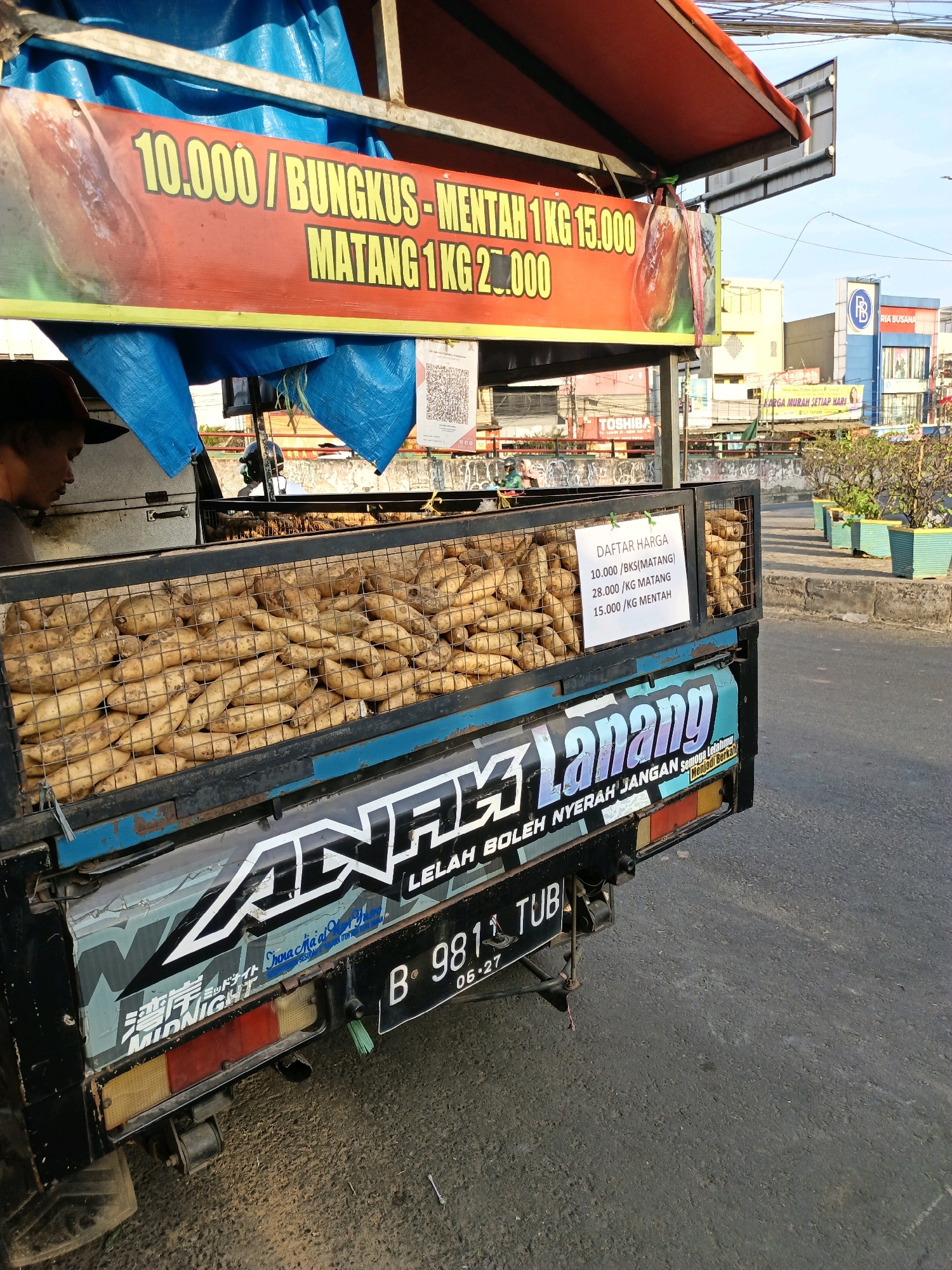 A street vendor sells sweet potatoes from a modified pickup truck in South Tangerang, Indonesia. The truck is adorned with colorful banners advertising prices: "10.000 / BUNGKUS - MENTAH 1 KG 15.000 MATANG 1 KG 25.000". A sign inside the truck displays a price list for sweet potatoes: "DAFTAR HARGA 10.000 / BKS (MATANG) 28.000 / KG MATANG 15.000 / KG MENTAH". The truck is loaded with sweet potatoes in a metal cage, and decorative stickers, including "ANAK LANANG" and "MIDNIGHT," are affixed to its sides. A person, possibly the vendor, is partially visible on the left, wearing a dark cap. The background shows a street scene with buildings, a traffic light, and potted plants. The sky is clear and blue, indicating daylight. The overall impression is of a busy street market in Indonesia.