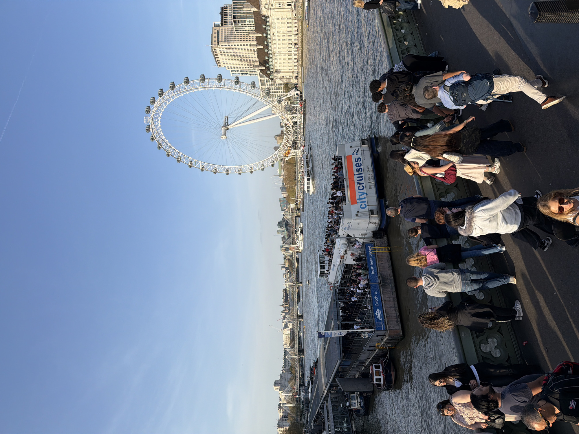 The image depicts a view in London, United Kingdom. The London Eye is prominently visible on the left. A cruise boat docks at a pier crowded with people boarding. A group of people are standing and walking on a bridge or elevated walkway overlooking the scene. The environment includes the River Thames, buildings along the riverbank, and a clear blue sky.