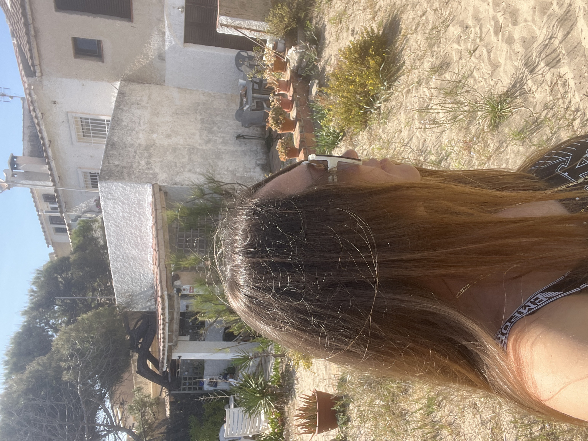 The image, taken outdoors in daylight, features a woman with long, wavy brown hair partially visible. The location appears to be La Marina De Creixell, Spain, evidenced by the style of residential buildings in the background. The background includes two buildings with white exteriors and brown window shutters. Several potted plants are arranged against one of the buildings. The ground is covered in sand with sparse vegetation. The woman is wearing sunglasses and a garment with the word "EMPORIO" visible. She seems to be either lying down or reclining, with her hair spread out on the sandy ground.