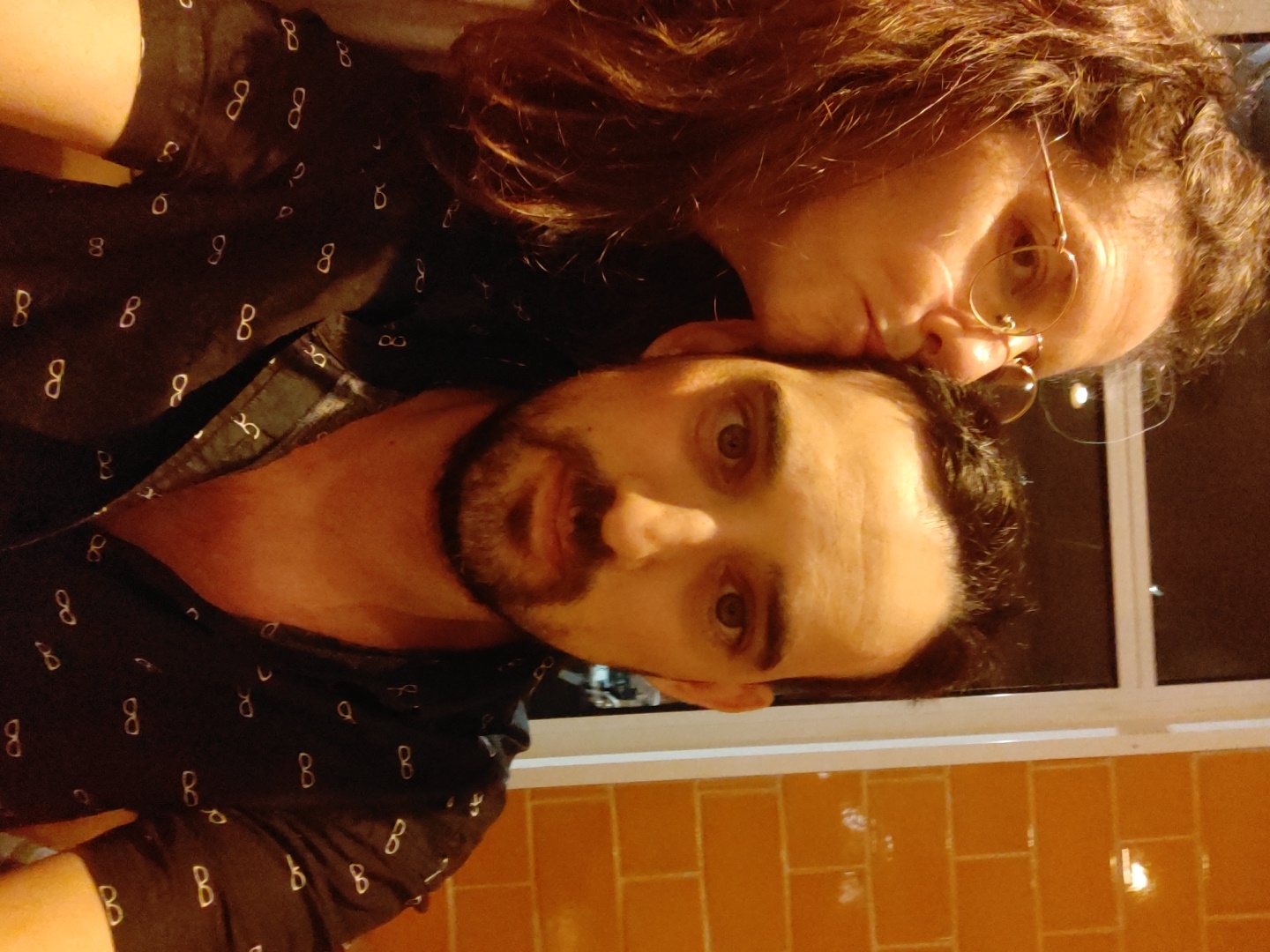 A man and woman pose indoors for a close-up photo. The man, with a beard, dark hair, and open shirt with a "number 8" pattern, looks directly at the camera. The woman, with curly brown hair and glasses, is kissing his forehead. The setting appears to be indoors, near a window and a tiled wall. There is no indication of the location in Tel-Aviv, Israel.