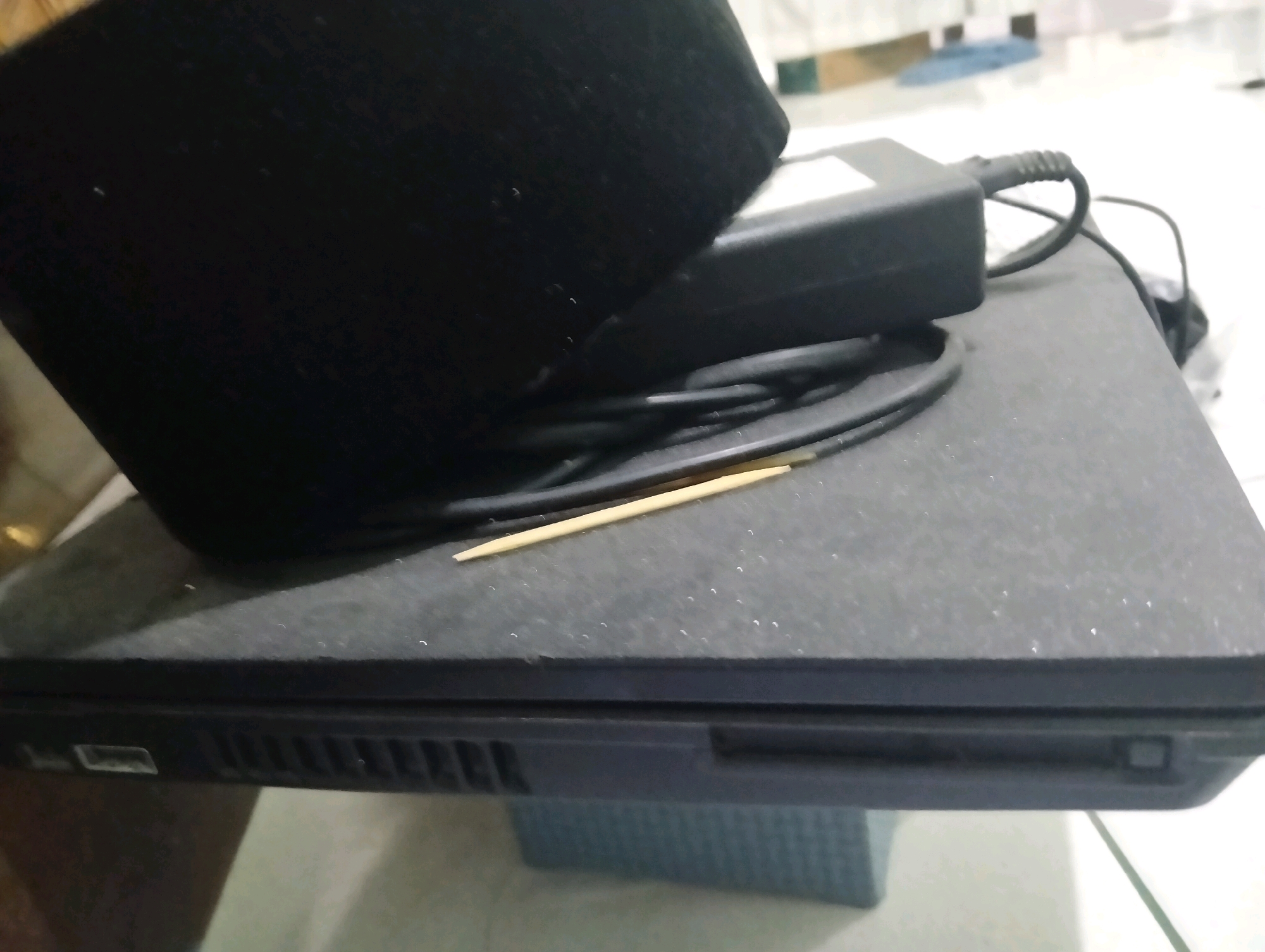 A black laptop is on a table. A black power adapter and its cord are on top of the laptop. A toothpick rests on the laptop's surface, near the center of the image. The laptop has ventilation grilles on its side, and a USB port is visible. The background is blurry, showing a light-colored surface and some indistinct objects. The overall scene appears to be indoors.