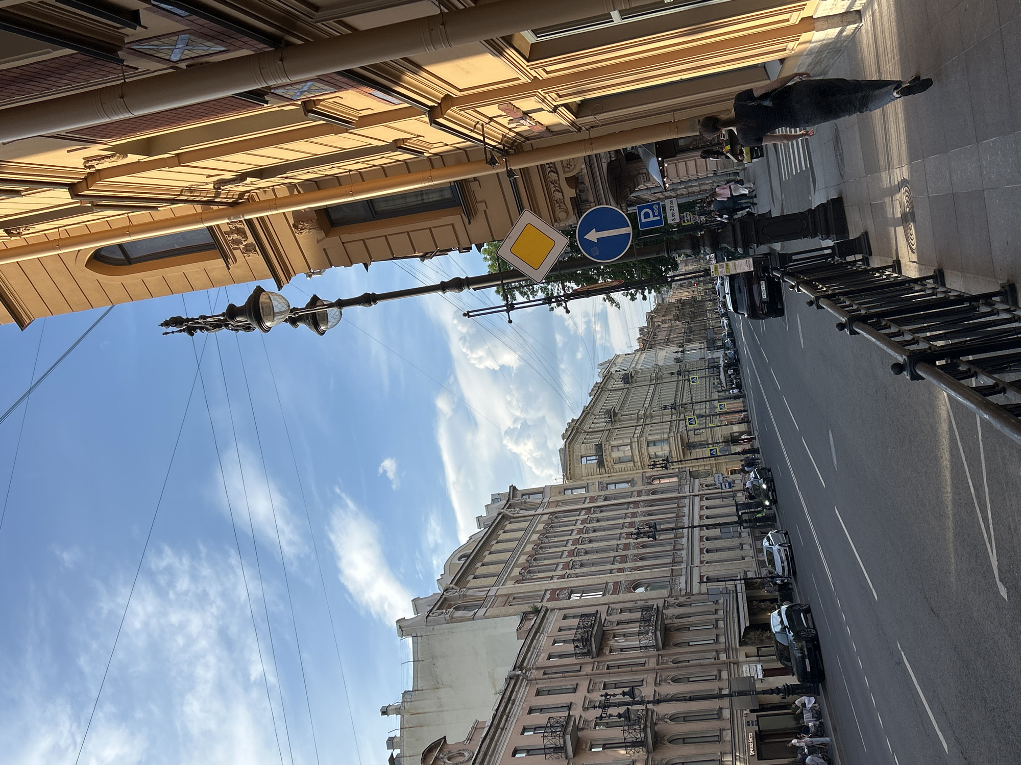 The image depicts a wide urban street in Saint Petersburg, Russia, under a bright blue sky with scattered white cumulus clouds, illuminated by the late afternoon or early evening sun. The perspective is from a paved sidewalk, looking down the street which recedes into the distance with a slight left curve.
On the immediate right, a black metal fence borders the sidewalk. Next to it, a prominent, multi-story building with an ornate, light-ochre facade stands, strongly illuminated by golden sunlight that highlights its architectural details and casts distinct shadows from its vertical drainpipes and window frames. A black, dual-globe streetlamp is positioned on the sidewalk in front of this building. Attached to the streetlamp post are two traffic signs: a yellow diamond with a white border (priority road) and a blue circular sign with a white upward arrow (one-way street). Below these, a blue square parking sign indicates "P 8.00-20.00". Further along this sidewalk, several pedestrians are visible, with one individual in dark clothing walking away from the camera in the foreground.
The asphalt road features multiple lanes demarcated by white dashed and solid lines. Several cars are parked along the left curb, including a black SUV with a Russian license plate "Y240YP 98" and a silver sedan. Other vehicles are present further down the street. Across the road, a continuous line of multi-story buildings, ranging in color from beige to pink, displays numerous windows, balconies, and decorative architectural elements. One establishment, labeled "греческое бистро" (Greek Bistro) in Cyrillic, has outdoor seating where several individuals are dining. Overhead, multiple dark utility wires span across the sky.