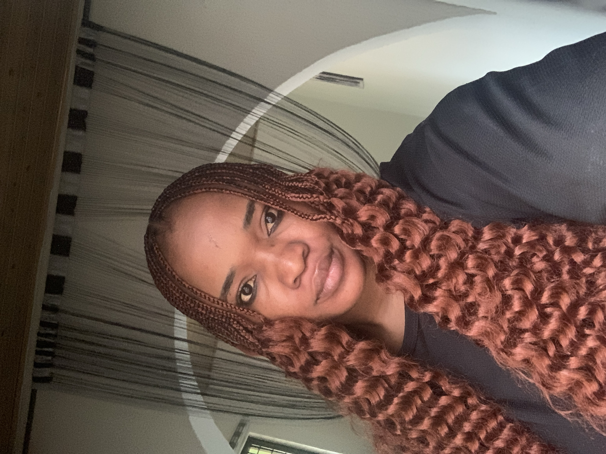 The image shows a woman with brown, braided hair and curly extensions, wearing a black shirt. She is positioned indoors, with a background featuring a textured curtain or screen and a light-colored ceiling with an air vent. The location, Barakin Akawo, Nigeria, is an unconfirmed detail as it's not discernible from the image itself.