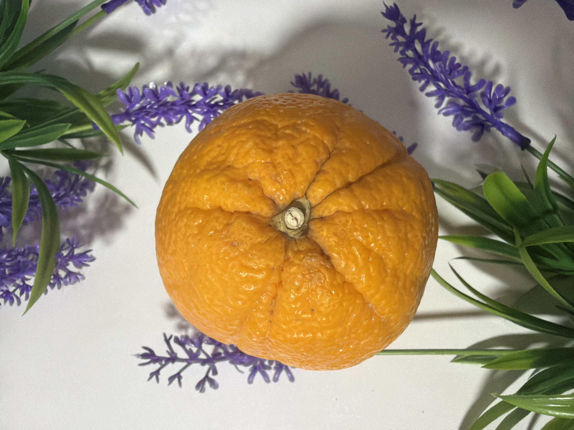 An orange sits in the center of the frame, surrounded by artificial purple lavender and green foliage on a white surface.