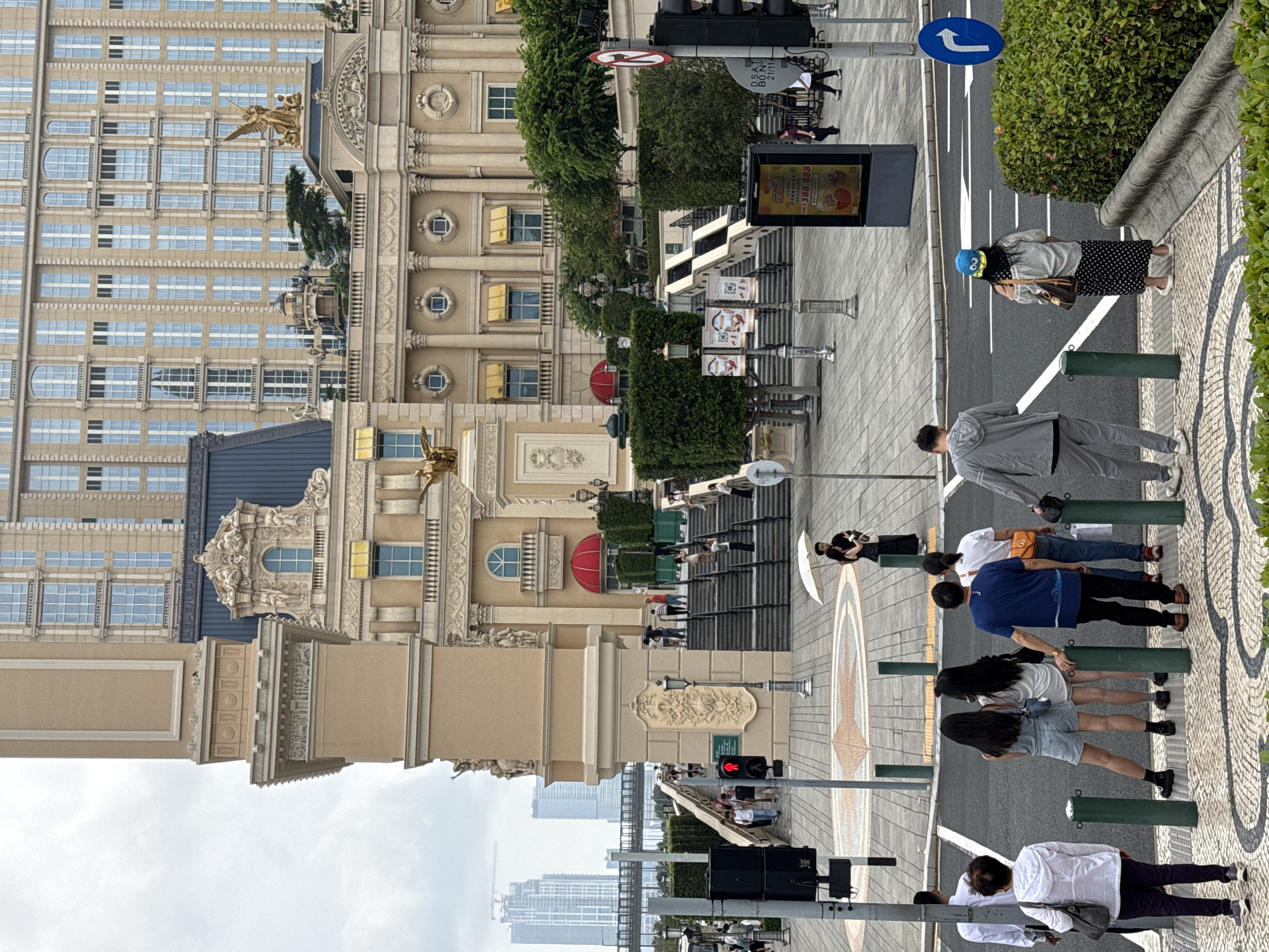 The image depicts a street scene in an urban environment. The architectural style of the buildings suggests a grand, possibly European-influenced design. The street is paved, with pedestrian crossings marked. Several pedestrians are visible, some waiting to cross the street at a traffic light. There are street signs and traffic signals present. The location is identified as Unknown, Macao.