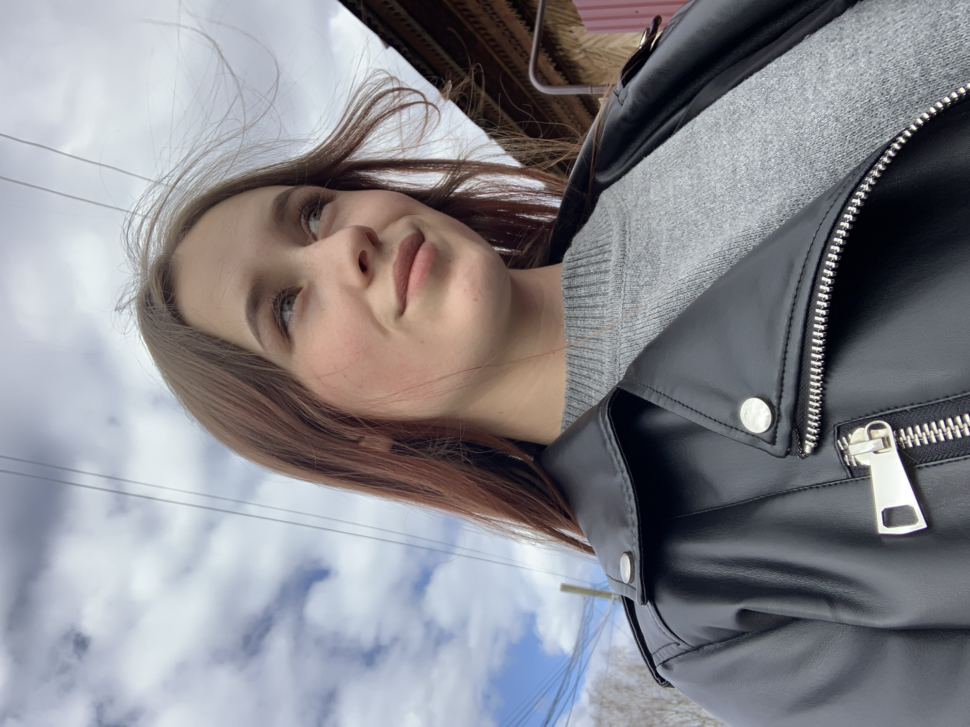 A young woman stands outdoors in Фоки, Russia. She is wearing a grey sweater and a black leather jacket. The sky behind her is cloudy with patches of blue. Wires are visible above her head. The background includes a wooden building structure.