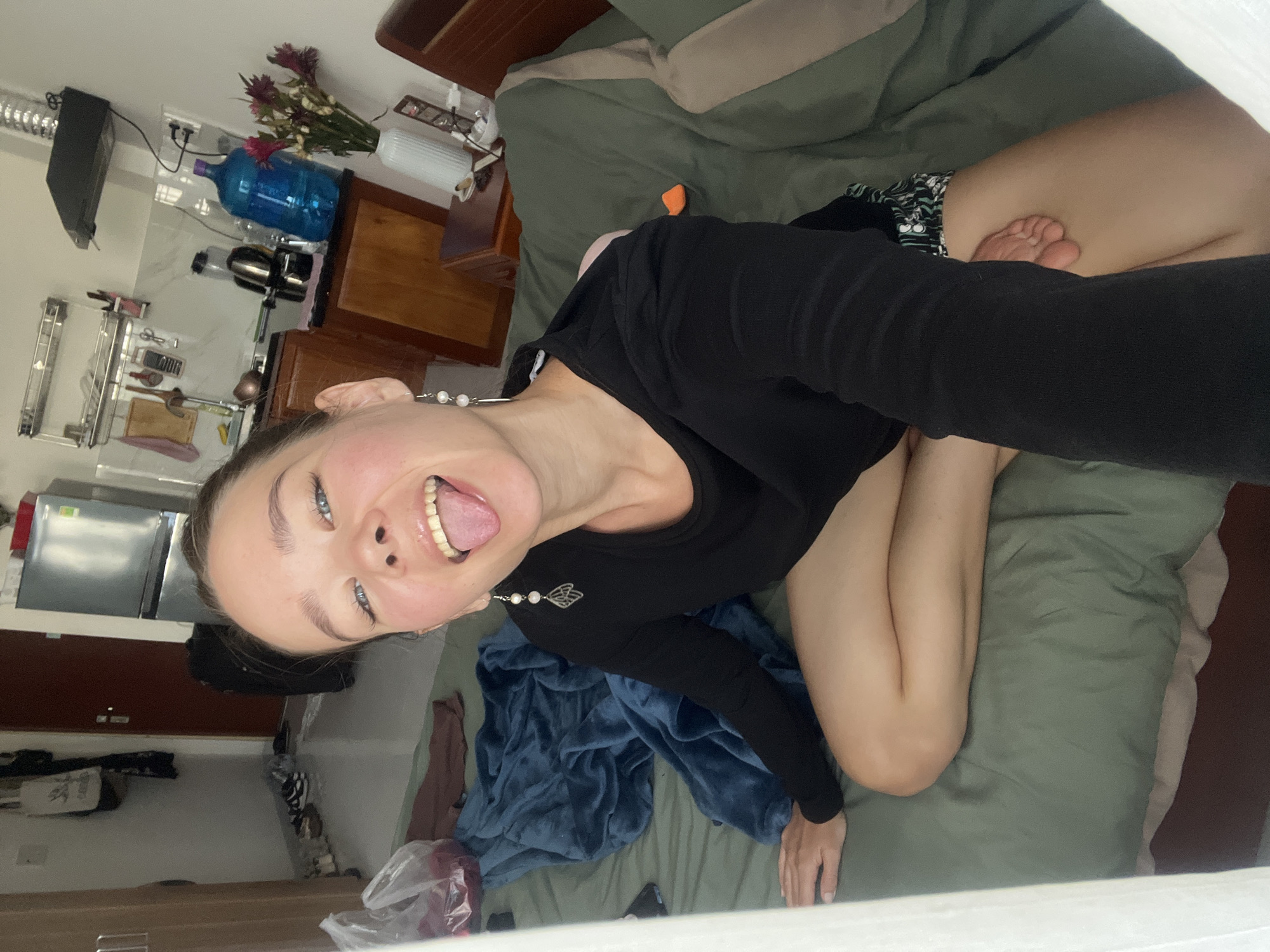 A young woman is positioned on a bed with green sheets, in what appears to be an apartment room in Nam Nha Trang Ward, Vietnam. She is wearing a black long-sleeved shirt and is kneeling on the bed. Her head is tilted, and she is sticking out her tongue. There is a kitchen area visible in the background with a refrigerator, cabinets, and various kitchen appliances. Other objects on the bed and around the room include a blue blanket, plastic bags, and furniture.