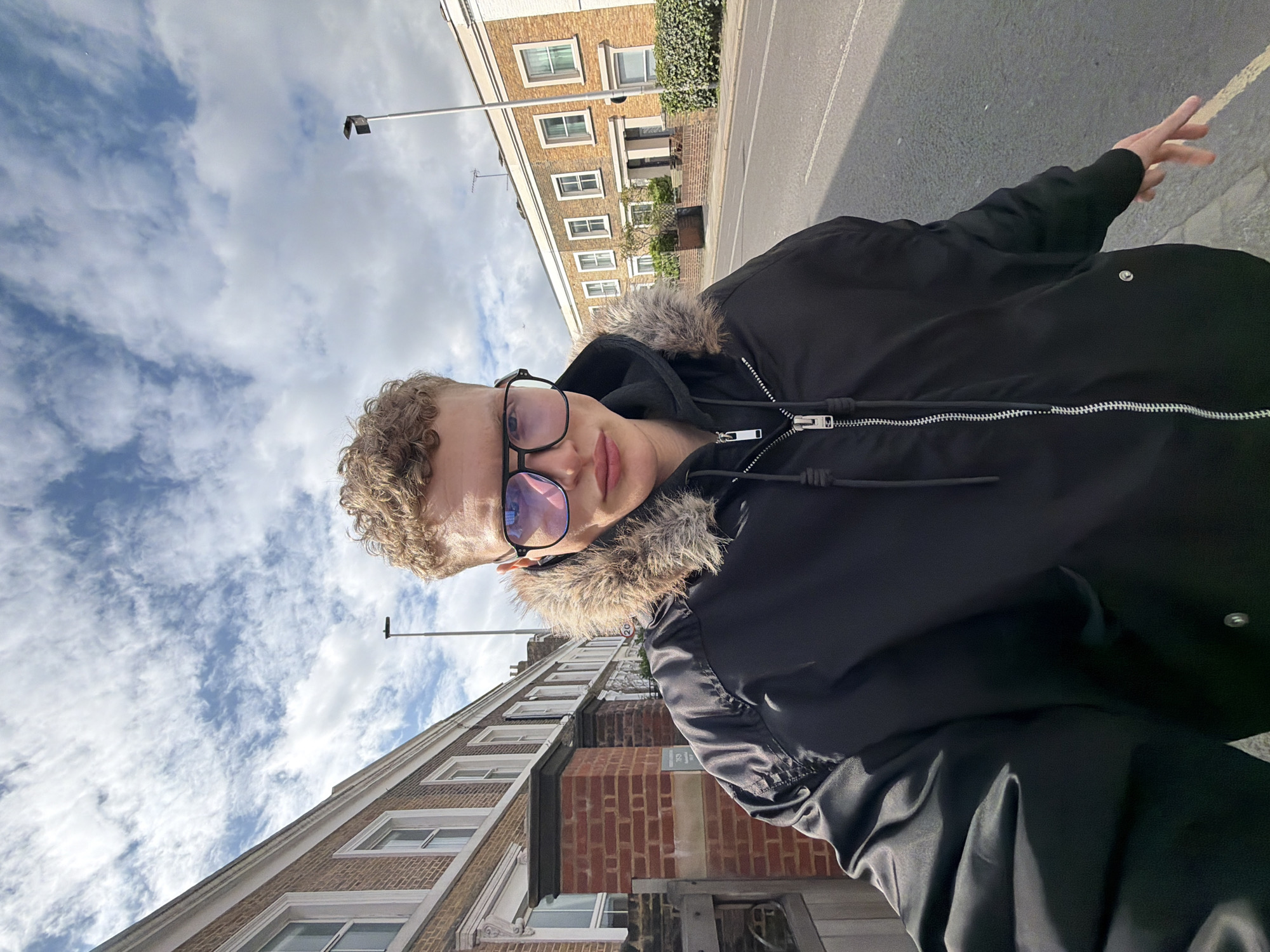 A person is taking a selfie outside on a street in London, United Kingdom. The person is wearing glasses and a black jacket with a fur-lined hood. The buildings are primarily brick with white window frames and are several stories tall. The sky is blue with scattered white clouds. A streetlight is visible in the background. The person's hand is partially visible on the right side of the frame. The road is made of asphalt.