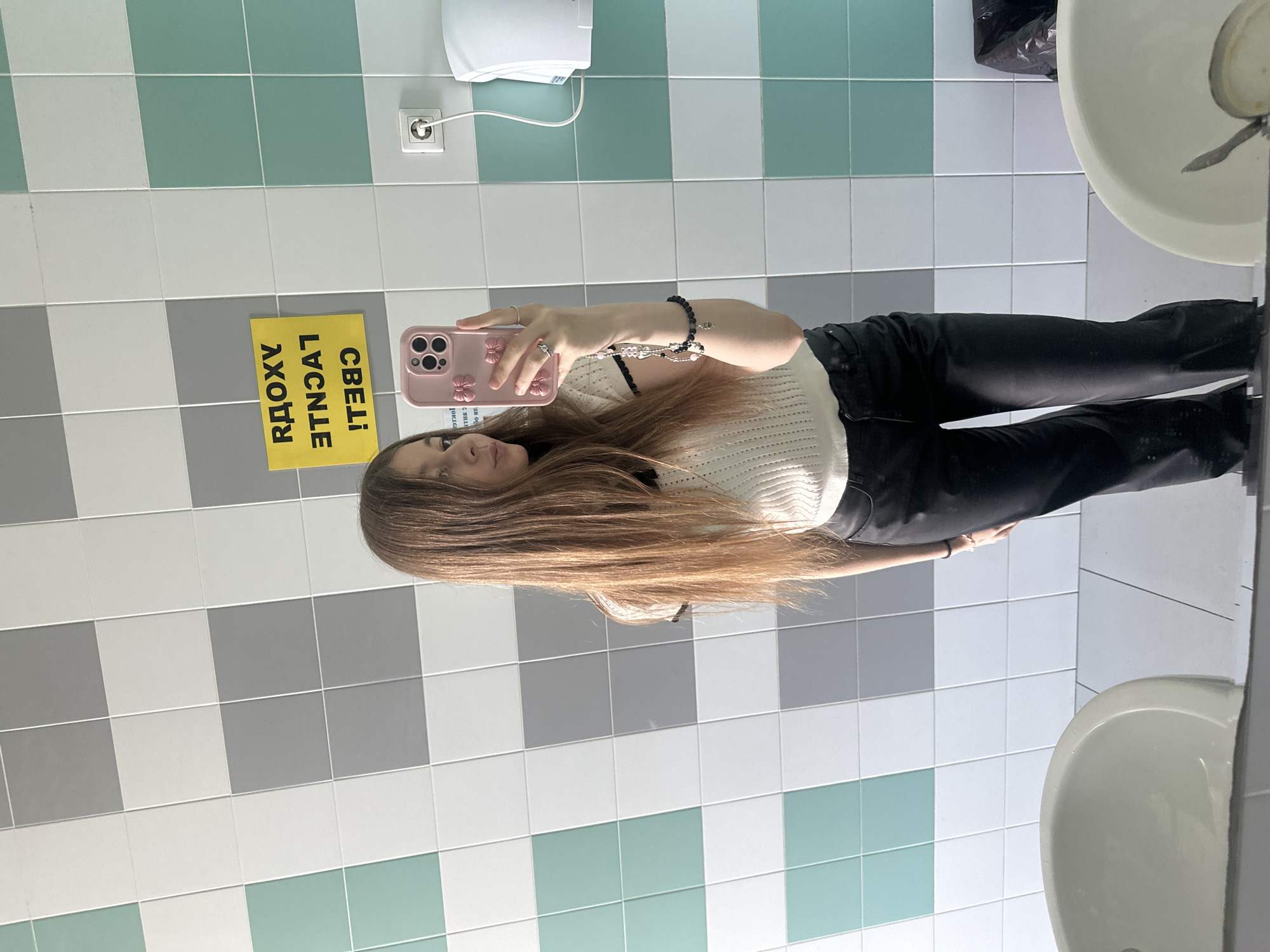 The image shows a young woman taking a selfie in a bathroom. She is holding a pink phone case. She is wearing a light-colored sweater and black pants. There are two sinks visible in the bathroom, along with tiled walls in a mix of green, white, and gray. A yellow sign is visible in the background with reversed Cyrillic text. Based on the reversed text "СВЕТ! ЛУЧШЕ! АДОХУ", the sign translates to "LIGHT! BETTER! A LOT!". The bathroom is located in Kazan, Russia.