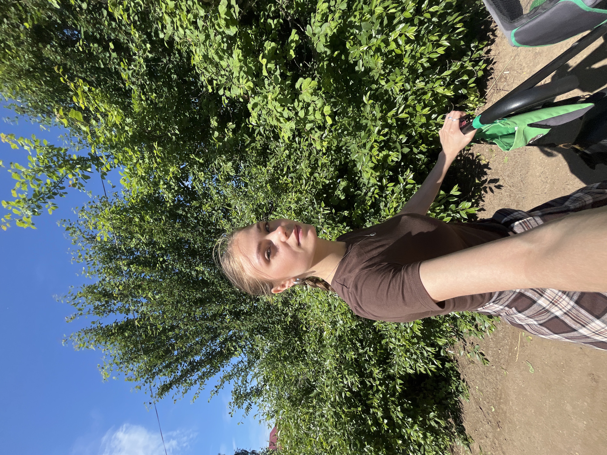 A young female, appearing to take a selfie, is positioned outdoors in Фоки, Russia, against a backdrop of dense green foliage and a clear blue sky. She has light brown hair pulled back into a braid on her left side and is wearing a plain brown short-sleeved t-shirt over a brown and beige plaid skirt or shorts. She wears silver hoop earrings, a silver necklace with a small pendant, and a stud piercing on her left nostril. Her left hand is gripping the black handle of a green stroller or pushchair, which has a green fabric accessory attached. A thin black wire runs horizontally across the upper left sky and through the trees. The ground beneath her is a reddish-brown dirt path. A small portion of a red roof is visible behind the foliage on the far left.