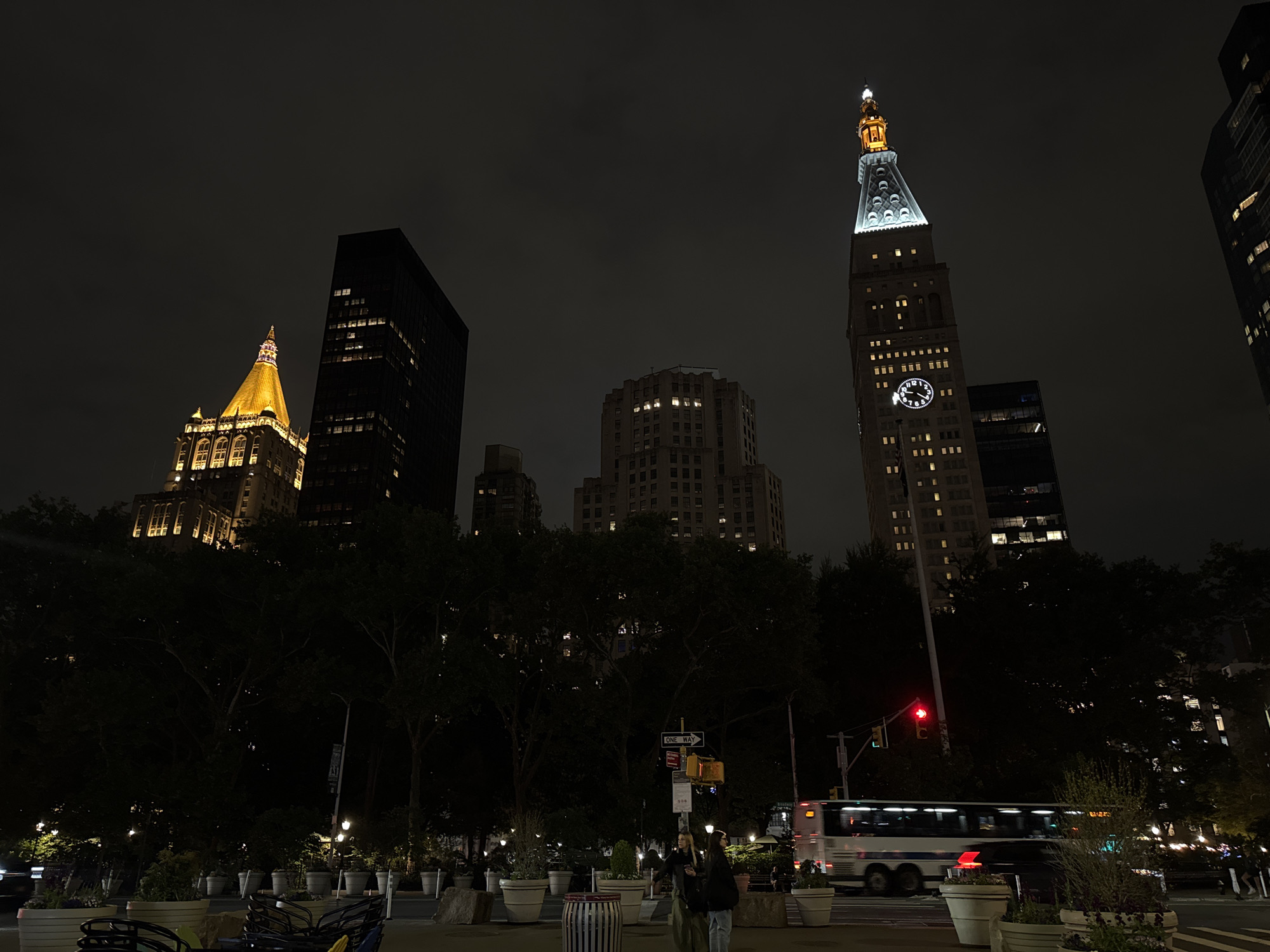 The image captures a nighttime scene in New York City, likely near a park, with several prominent skyscrapers in the background. From left to right, the buildings include a structure with a golden, ornate dome-shaped top, a dark, rectangular skyscraper, a light-colored building with numerous windows, and the iconic clock tower of One Madison Avenue, distinguished by its white clock faces and illuminated spire.
In the foreground, there are planters, trees, and benches suggesting a park or public space. People are seen walking along the sidewalk. A large bus is moving through an intersection where a traffic light is visible. Street signs and lamp posts are present among the foliage, adding to the urban atmosphere. The overall mood is dark and illuminated only by street lights and building lights.