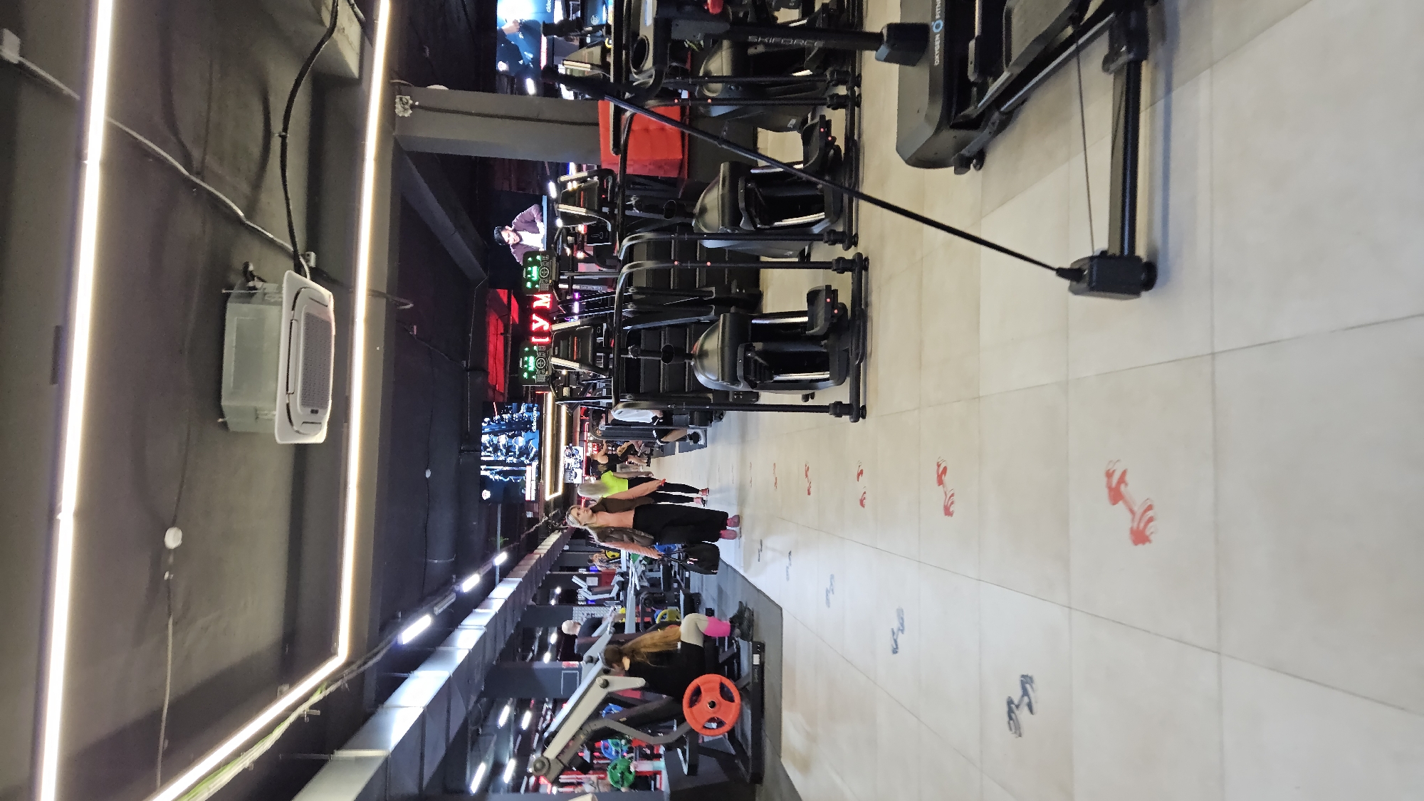 The image depicts an indoor fitness facility. Several rows of exercise equipment, including rowing machines and weight machines, are visible. People are exercising on the equipment. The walls are dark, with bright vertical light fixtures. Red dumbbell icons are painted on the floor. An air conditioning unit is mounted on the wall. The location and context of the image within Sevastopol, Ukraine are not discernible from the provided visual information.