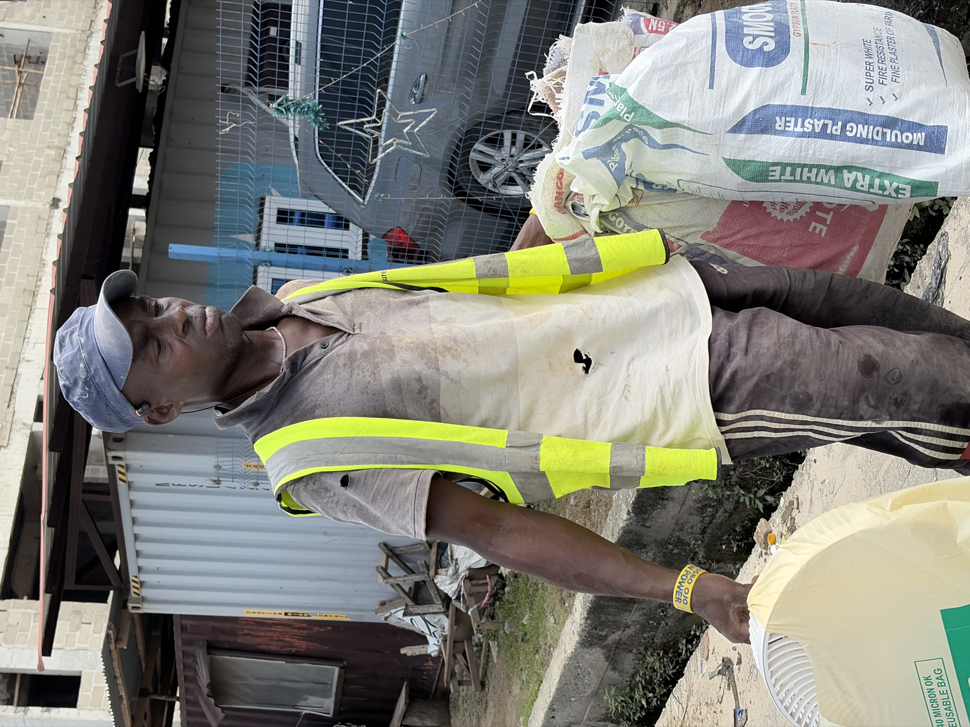 The image shows a man in Igboefon, Nigeria, wearing a dirty gray shirt, dark pants with white stripes, a safety vest, and a blue cap. He is carrying a white bag of "SMOR GYPSUM PLASTER" and another bag labeled "40 MICRON OK REUSABLE BAG". He wears a yellow wristband with the inscription "NGBO OJO OW POWER". Behind him is a metal fence with a car parked behind it. In the background, there's a building under construction and a shipping container. The environment appears to be a construction or storage area.