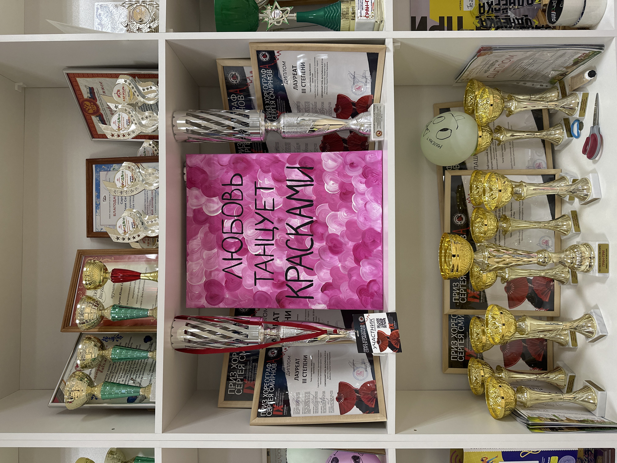 A well-organized shelf displays a collection of trophies, awards, and various items. The shelf is divided into compartments, each containing unique elements. The left compartment features an array of trophies and framed certificates, some with Russian text. In the center compartment, a pink book with Cyrillic script titled "Любовь Танцует Красками" ("Love Dances with Paints") is positioned between silver trophies and framed awards. The right compartment contains golden trophies, framed certificates, a deflated balloon with a drawn face, and small objects like scissors. The presence of multiple awards, certificates, and trophies indicates an achievement-oriented setting, likely related to dance or performance arts, possibly from an event or competition held in Votkinsk, Russia, as indicated by some awards.
