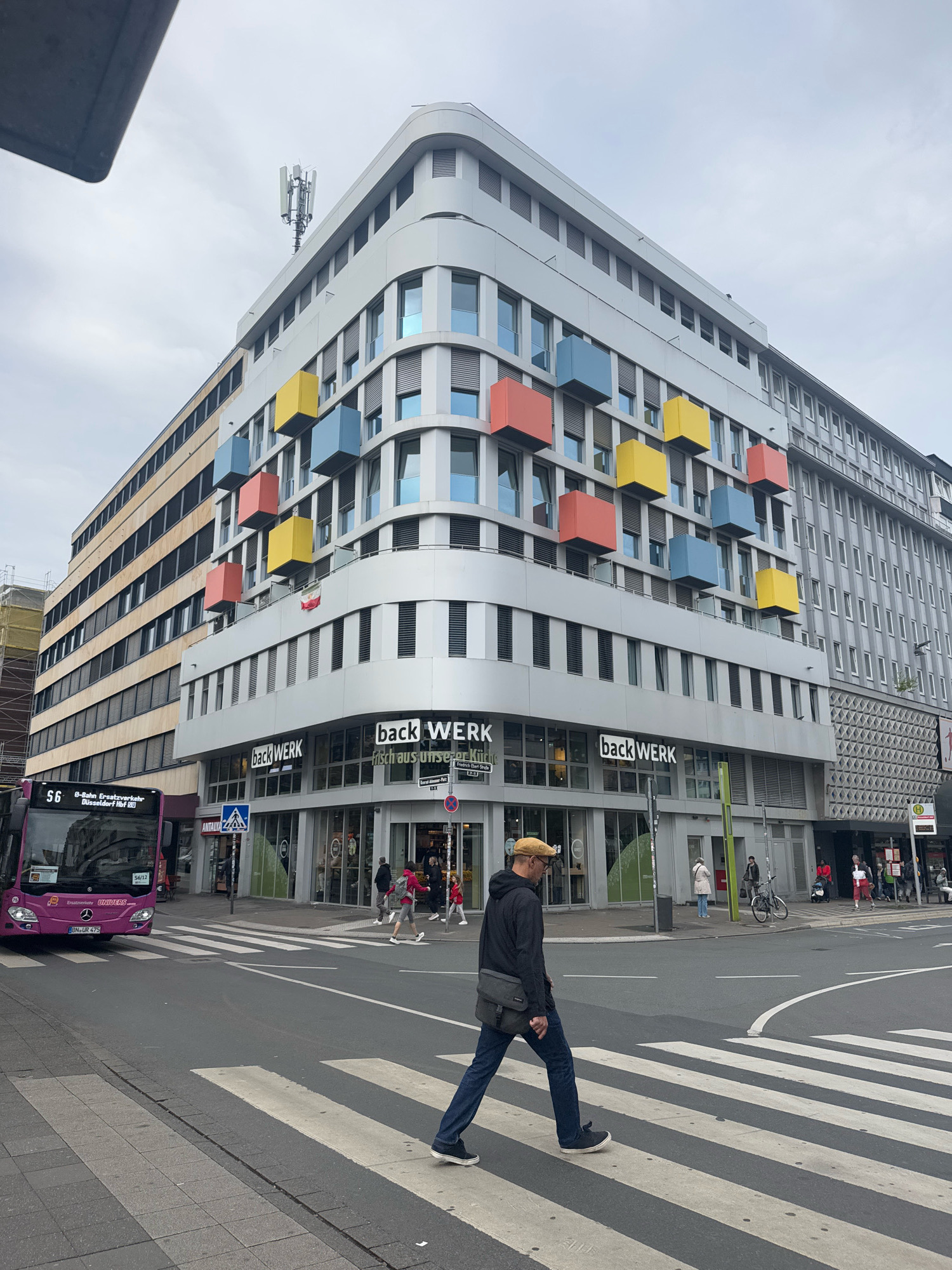 The image shows a street scene in Dusseldorf, Germany, likely near the Konigsallee given the architecture and the presence of a public transportation bus. A modern building with colorful geometric protrusions is prominently featured on the left. The building has a facade with a grid of windows and is adorned with yellow, blue, and red cubes that extend outwards. A bus, labeled "56 S-Bahn Ersatzverkehr Düsseldorf Hbf OO", is stopped at a crosswalk on the left side of the frame. In the foreground, a man wearing a yellow cap and a dark jacket is walking across the crosswalk. Several other pedestrians are visible in the background, some near storefronts. The environment is urban, with a street, sidewalks, and other buildings visible. The sky is overcast.