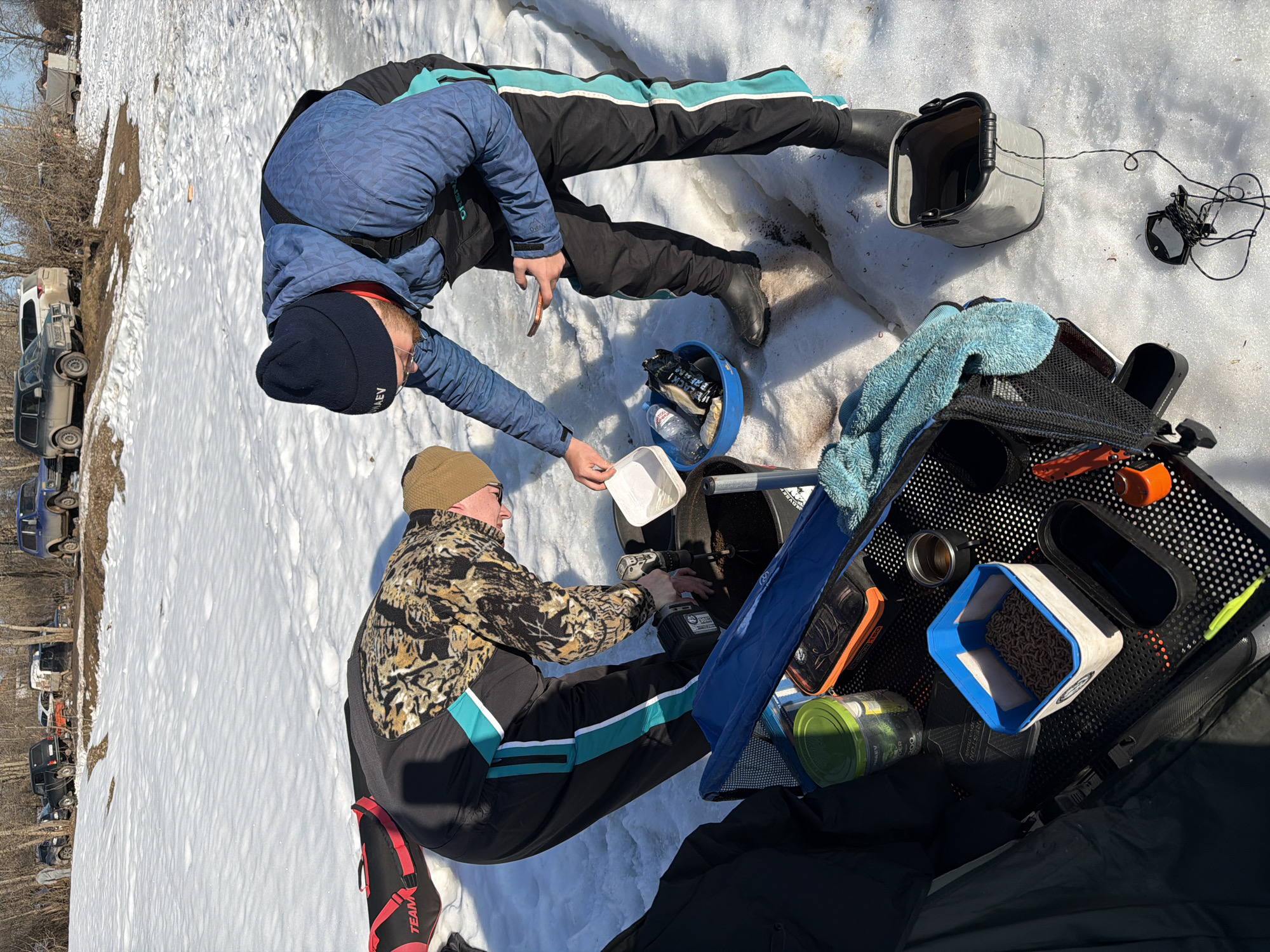 Two individuals are ice fishing on a snow-covered surface in Chaikovsky, Russia. One person, wearing a camouflage jacket and a beanie, is drilling into the ice with a power drill. The second person, dressed in a blue jacket and a dark beanie, is handing the first person a white container. A black fishing crate sits nearby, containing various items including a container of bait. Fishing equipment, including a tip-up and a tackle box, is scattered around the area. A line of vehicles is parked in the background. The weather appears to be sunny.