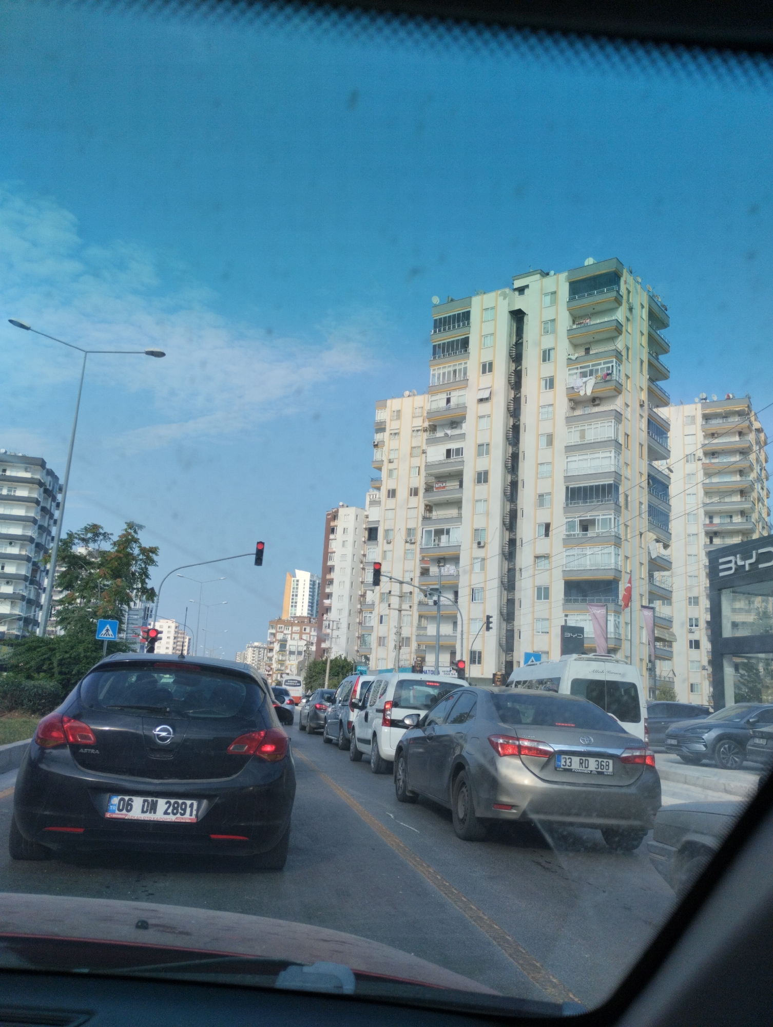 The image captures a street scene in Mezitli, Turkey, likely during daylight hours due to the bright sky and clear visibility. The perspective is from inside a vehicle, suggesting it is a roadside view. The foreground is dominated by the hood and windshield of the car from which the photo is taken, with a portion of the dashboard visible.
The main focus of the image is a line of cars in traffic, positioned on a multi-lane road. The most prominent vehicle is a black Opel Astra in the left lane, with its license plate "06 DN 2891" clearly visible. To its right, a silver Toyota Corolla with the license plate "33 RD 368" is also in view, along with other vehicles further down the road. The traffic appears to be at a standstill or moving very slowly, indicated by the density of cars.
Towering apartment buildings line the right side of the street, characterized by their modern, multi-story architecture with balconies and numerous windows. The buildings are predominantly light-colored, with some featuring darker accents. Traffic lights are visible, with at least one showing a red signal, which likely contributes to the traffic congestion. Streetlights and utility poles are also present along the roadside.
Vegetation, including trees and bushes, can be seen on the left side of the road, providing a contrast to the urban structures. The sky is a clear blue with some faint clouds. The overall impression is that of a typical urban traffic situation in a populated area.