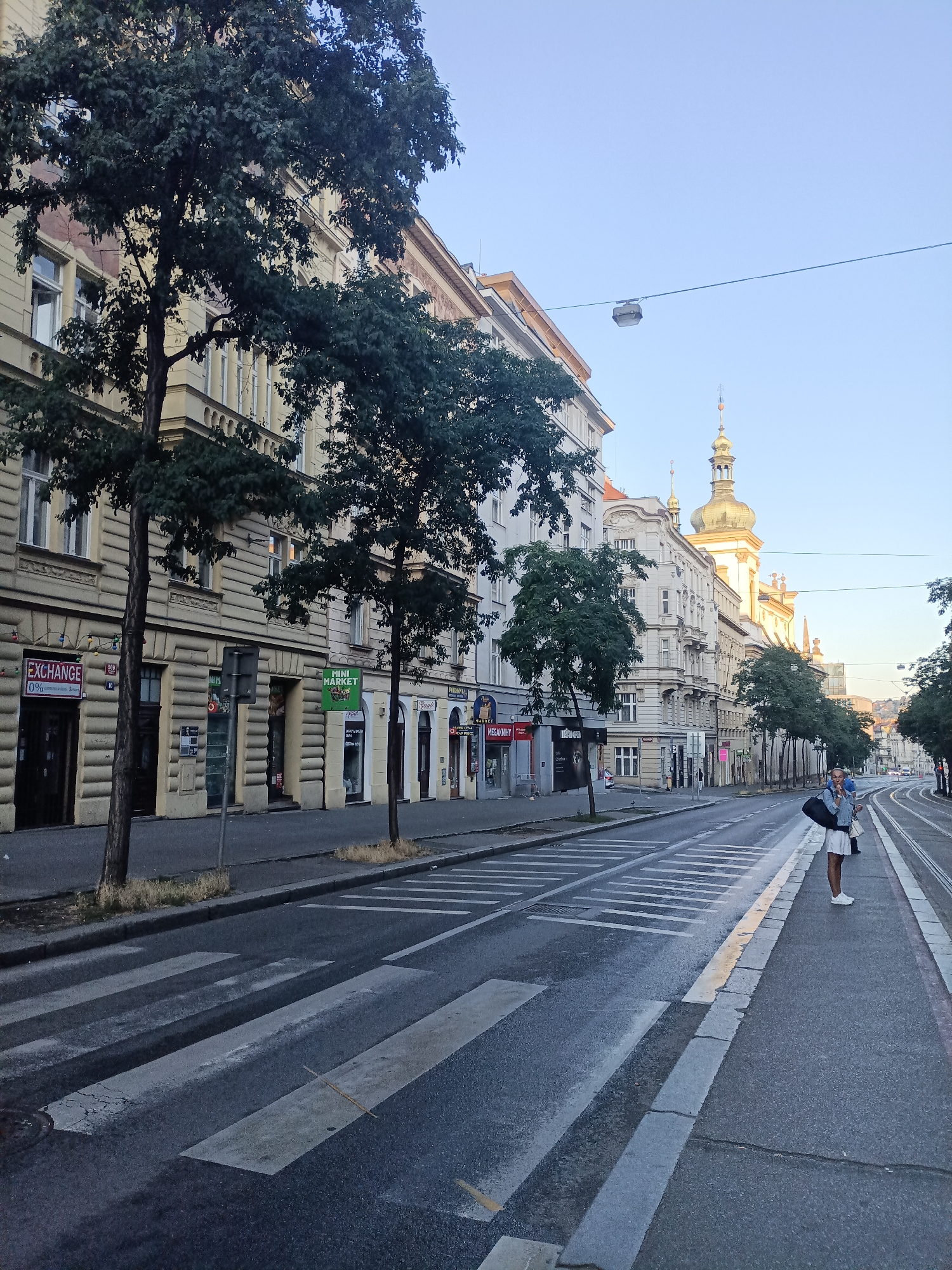 The image depicts a street scene in Prague, Czechia, likely during the daytime with clear skies. A wide street runs diagonally from the foreground to the background, featuring tram tracks and a pedestrian crossing in the foreground. Buildings line the left side of the street, exhibiting architectural styles typical of European cities, with multiple stories and decorative facades. Trees with green foliage are interspersed along the sidewalk. On the right side of the street, a single person, a woman, stands near the tram tracks, holding a bag over her shoulder and looking towards the left. She is wearing a light-colored skirt and a denim jacket. In the distance, more buildings are visible, including one with a prominent dome and spire. The street appears to be a mixed-use area, with shops and commercial establishments visible on the ground floors of the buildings. The overall impression is one of an urban environment during a calm period.