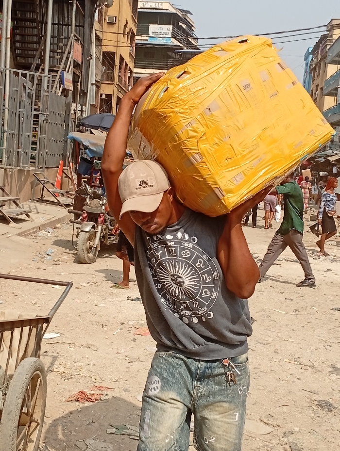 A man carries a large, yellow-taped box on his shoulders in a bustling, urban environment. He wears a light-colored baseball cap, a gray t-shirt with a zodiac design, and denim shorts with faded writing. A metal cart with a wheel is visible to his left. The backdrop features buildings of varying heights and architectural styles, while other people are walking in the background, along with a motorcycle and an umbrella. The ground appears dusty and scattered with debris.