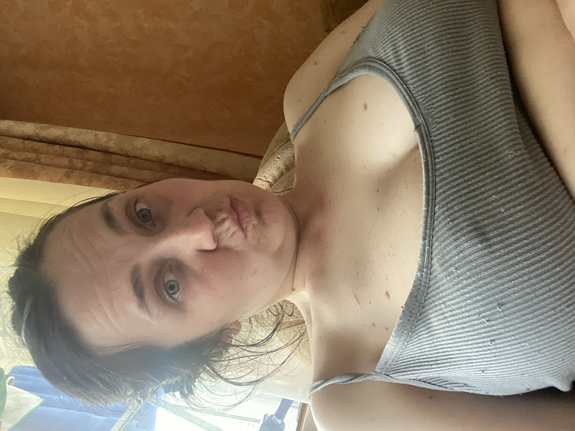 This is a close-up, vertically oriented selfie of a young woman. She is looking directly at the camera with her lips pursed. Her eyes are wide open and appear to be blue or green. She has dark hair that is slightly wet and appears to be pulled back loosely. She is wearing a gray ribbed tank top that reveals her shoulder and part of her chest. There are several small moles visible on her skin.
The background is out of focus, but suggests an indoor setting. To the left, there is a textured orange wall or surface, possibly wallpaper or a painted wall. Below that, a lighter colored fabric is visible, possibly curtains or blinds, with horizontal folds. The lighting suggests it is daytime, possibly afternoon, with natural light coming from the right side of the frame. The overall mood seems casual and playful, suggested by the woman's facial expression. There is no visible text in the image.