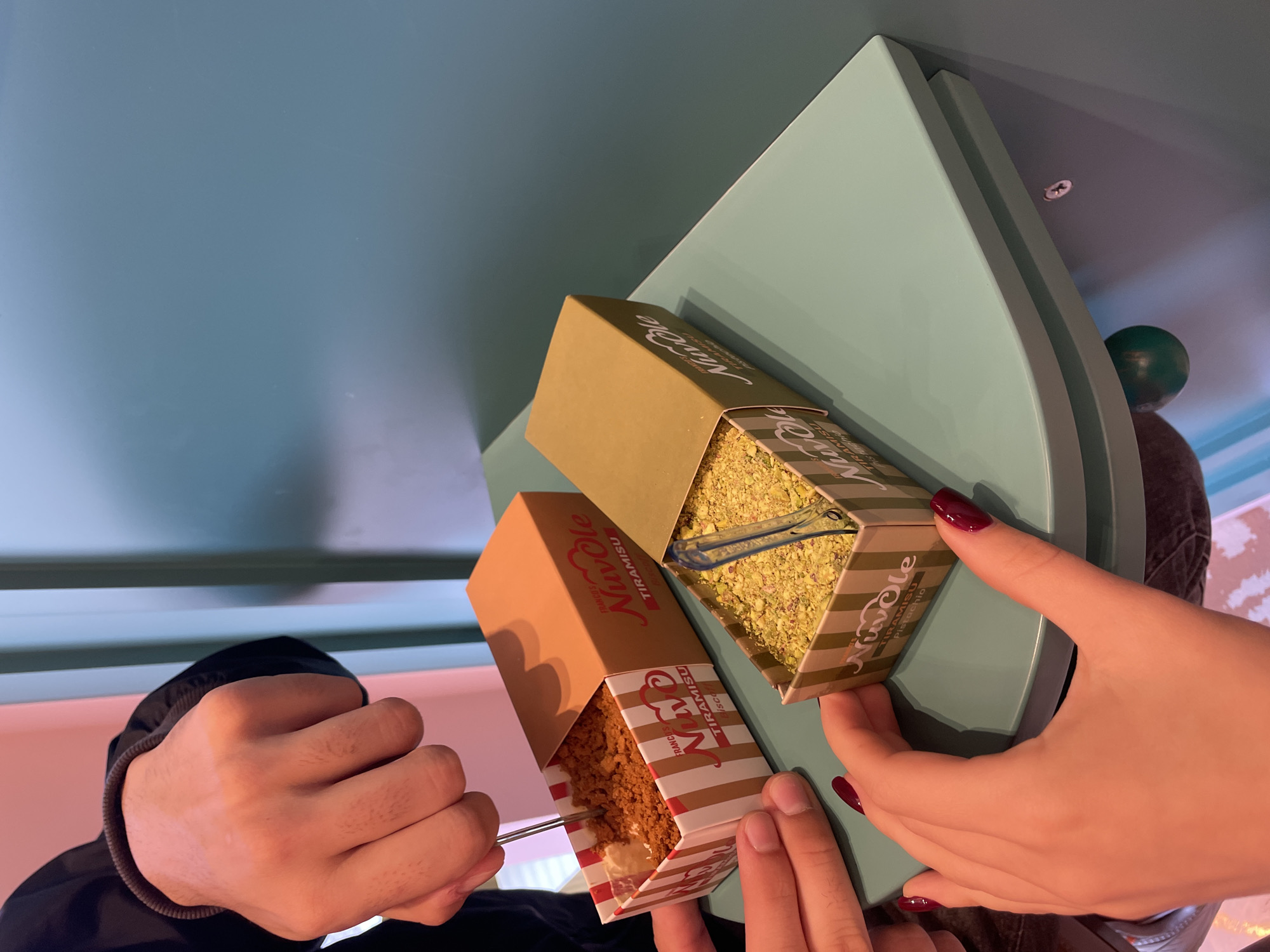 Two people are holding takeaway boxes of "Nuvole Tiramisu Pistachio" over a light-blue surface. One person is wearing a black jacket and is eating the tiramisu from the box with a fork. The other person is holding the other box and has their nails painted a dark red color. The background appears to be inside a building, possibly a cafe or restaurant. It is likely in Sofia, Bulgaria as "Nuvole" tiramisu is a brand that exists there.