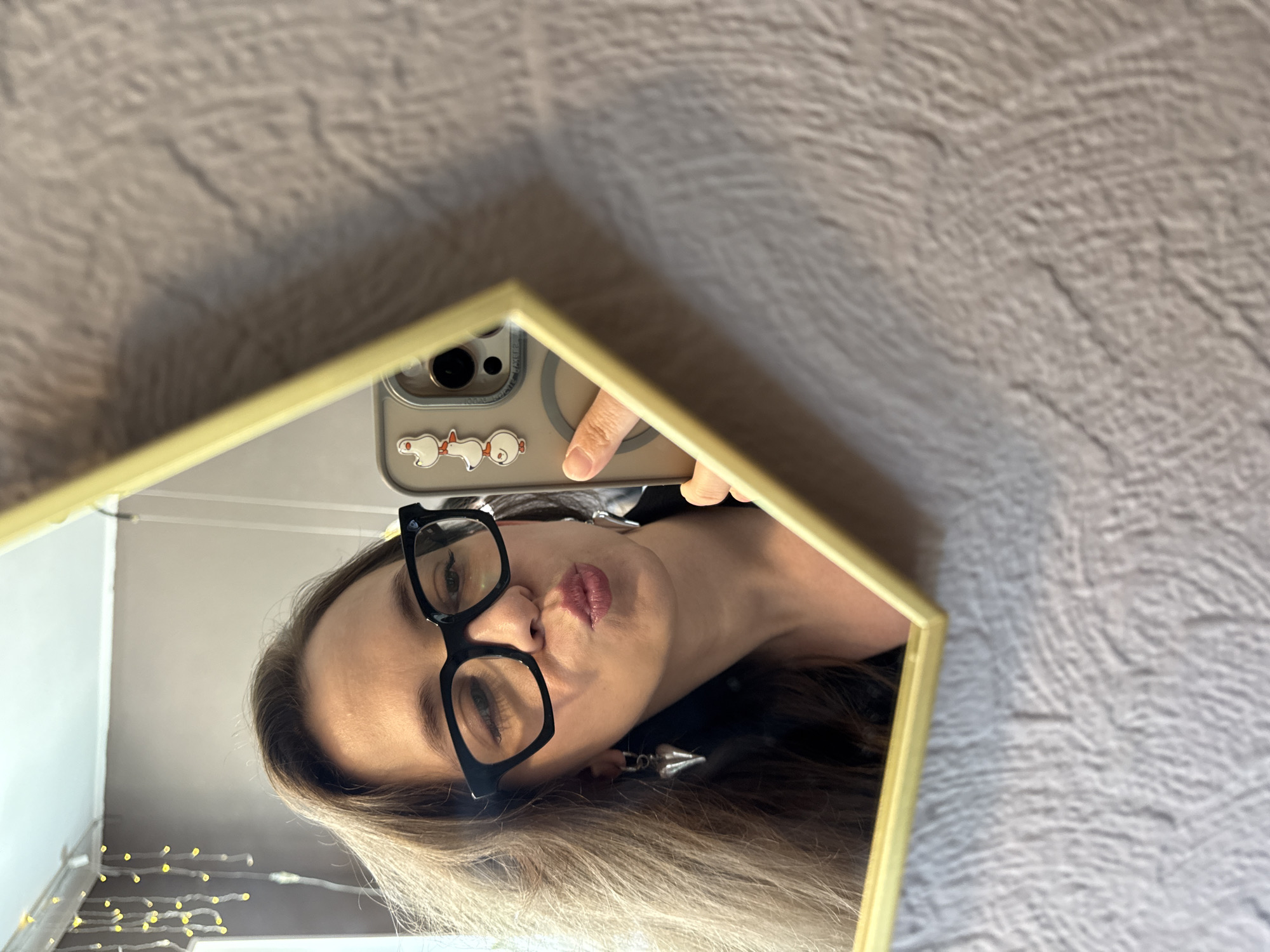A woman is taking a mirror selfie in a room. She is wearing black-rimmed glasses and has long, brown hair. Her lips are puckered as she holds up a phone with a case decorated with duck stickers. She is wearing silver earrings in the shape of hearts. The mirror she is using is hexagonal and has a gold frame. The background of the room is blurred, but shows a wall with a textured pattern and string lights with yellow bulbs hanging from the ceiling. The image was taken in Chaikovsky, Russia.