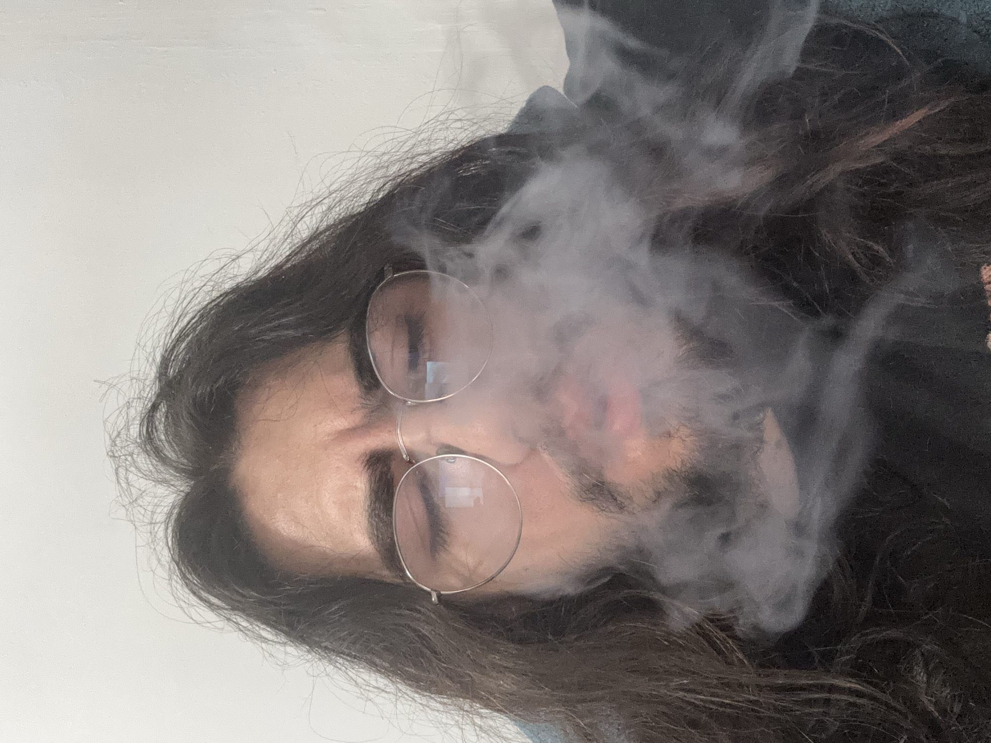 A close-up, eye-level, medium shot shows a person exhaling a cloud of white smoke. The person, who has long, dark hair and is wearing round, thin-rimmed glasses, is looking down. Their eyebrows are furrowed. The background is a plain, textured, off-white wall.