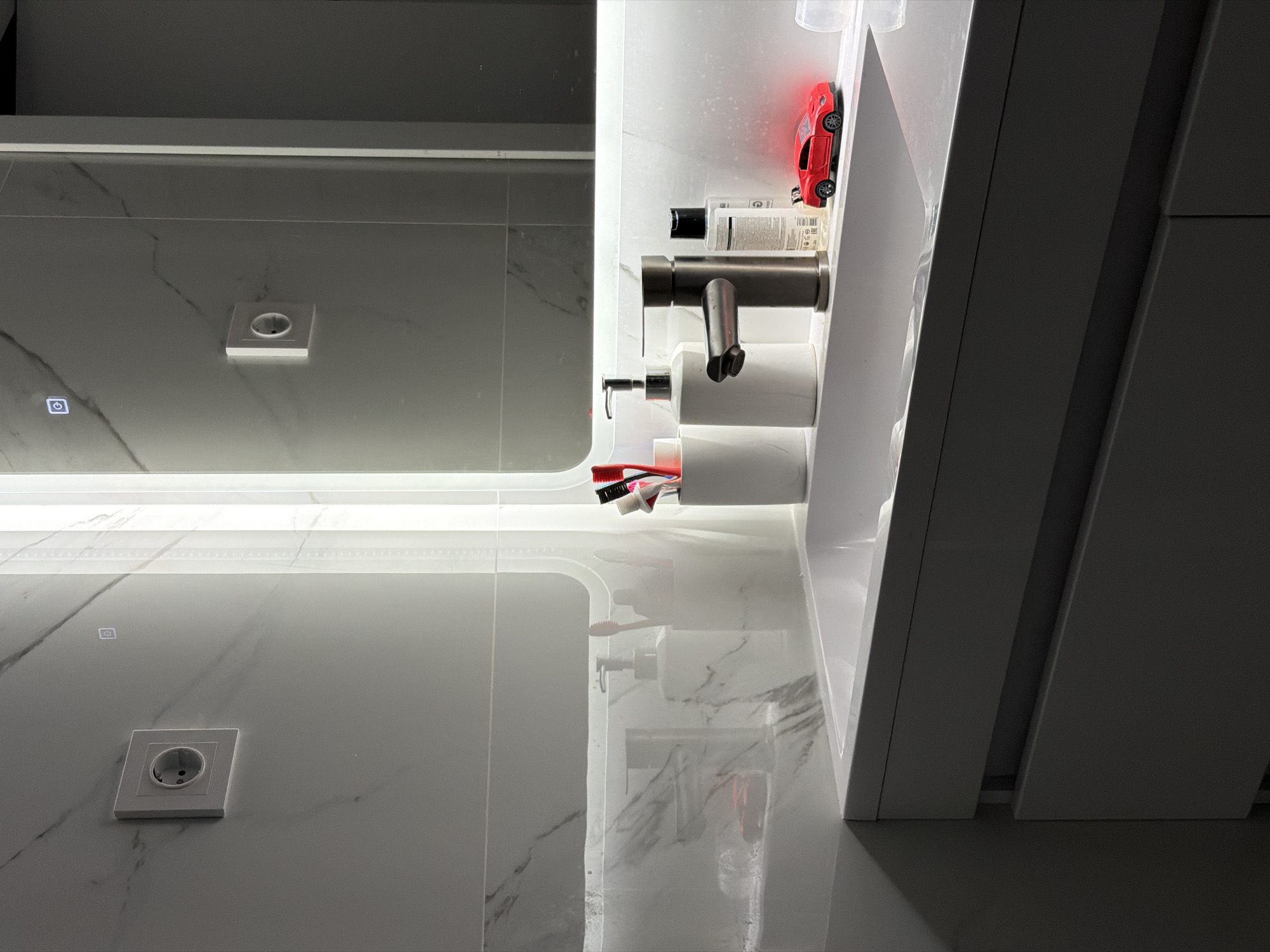 The image shows a bathroom interior. The walls are white with gray marble veining. A mirror with integrated lighting reflects the wall on the opposite side. The mirror also features a small touch-sensitive button, likely for controlling the light. On the right side of the image, an open cabinet reveals toiletries, a silver faucet, a container holding toothbrushes, and a small red toy car. The cabinet's door is white and blends with the adjacent wall. An electrical outlet is visible on the wall, both directly and in the mirror's reflection. The location is Chaikovsky, Russia.