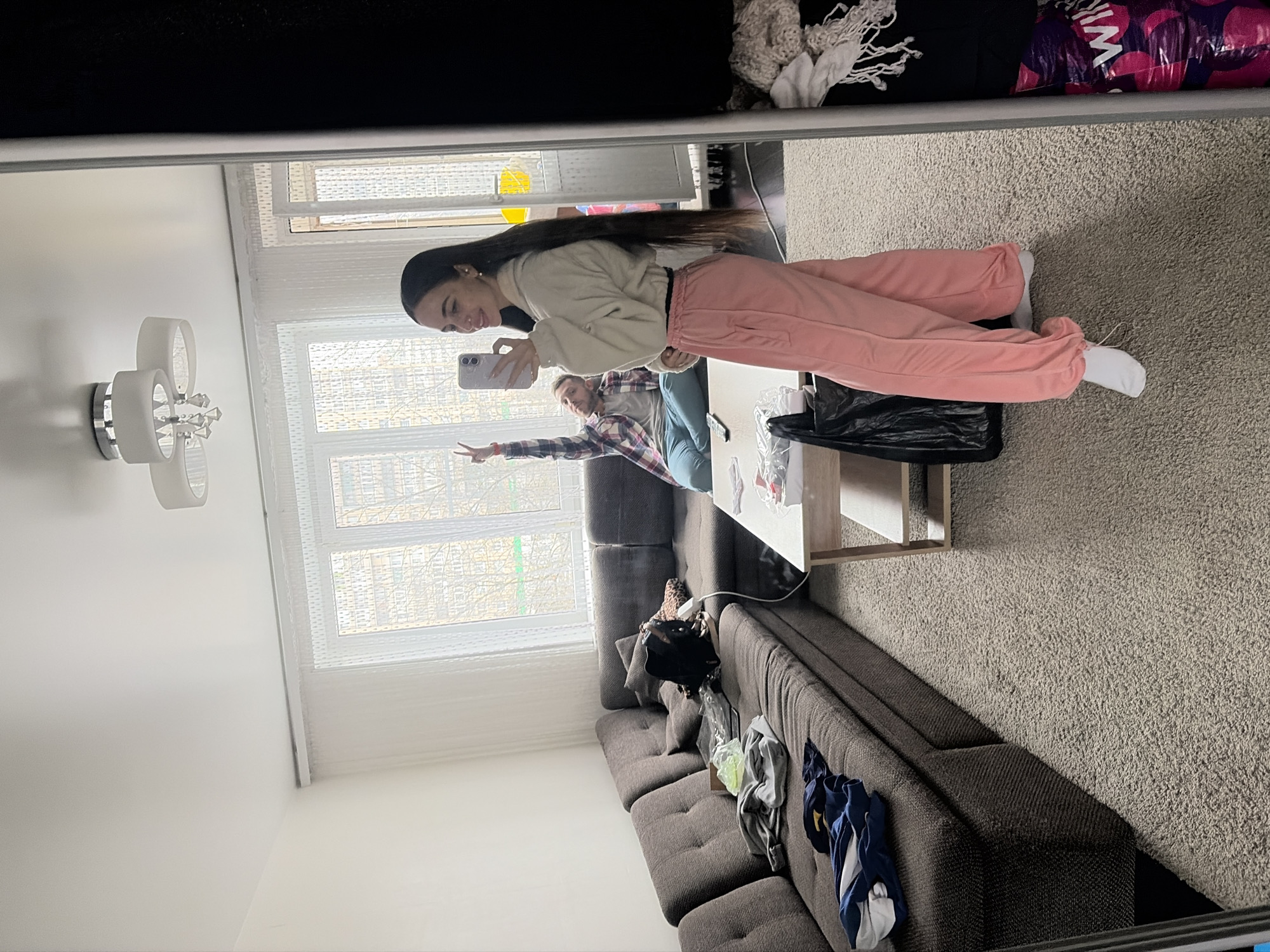 A reflection in a mirror captures a residential interior. A young woman with long, dark hair, wearing a beige sweatshirt and pink sweatpants, holds a smartphone and appears to be taking a selfie. In the background, visible through the mirror, another person is seated on a grey couch and raises their hand in a V-sign. The room contains a window, a light fixture, and a coffee table with items on it. The visible items on the couch include clothing and accessories. Based on the style and architecture visible through the window, the location could be a residential area in Saint Petersburg, Russia. The environment is a mix of interior living space with a view of an external cityscape.
