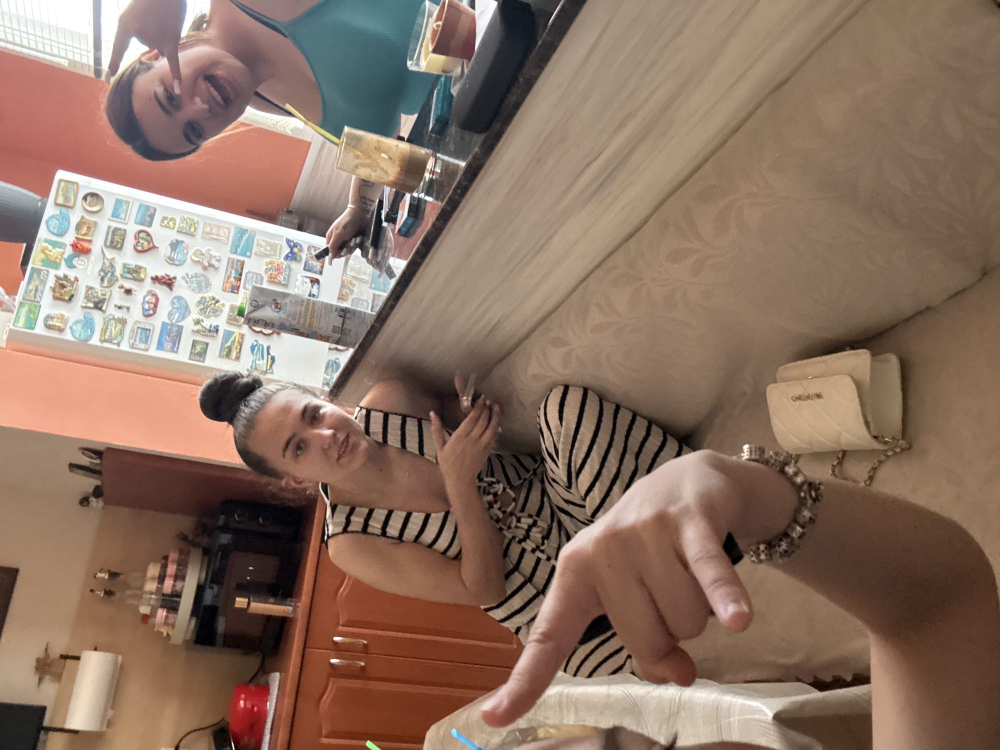 This media file captures a casual indoor scene featuring two young women, likely in a home kitchen or combined living space in Kyustendil, Bulgaria. The image is rotated 90 degrees counter-clockwise.
The primary subjects are two women. In the upper part of the frame (when corrected), a woman with dark hair tied back is visible, wearing a teal-colored top. She is smiling broadly and making a peace sign towards the camera. On a lower level, another woman is seated, wearing a black and white striped jumpsuit or dress, with her hair in a bun. She looks towards the camera with a subtle smile. Her left hand is raised towards her chest, while a hand belonging to the person holding the camera is prominently displayed in the foreground, adorned with a beaded bracelet and pointing downwards.
The setting appears to be a cozy domestic environment. A light-colored sofa or bench with an ornate pattern is visible on the right. In the background, a light orange wall provides a warm tone. Kitchen elements include wooden cabinets of a similar orange-brown hue, a refrigerator heavily decorated with various souvenir magnets, and a paper towel holder. On a counter, there are two glasses with colorful drinks and straws, as well as a multi-tiered dessert stand.
The women appear to be relaxing and enjoying drinks, likely posing for a casual photo or selfie. Both exhibit cheerful and relaxed expressions. A notable detail is the small, white quilted handbag with a gold chain strap resting on the sofa, featuring the faintly visible text "GIORGIO ARMANI" on its clasp. The time of day is not explicitly clear but suggests a casual daytime or evening gathering indoors.