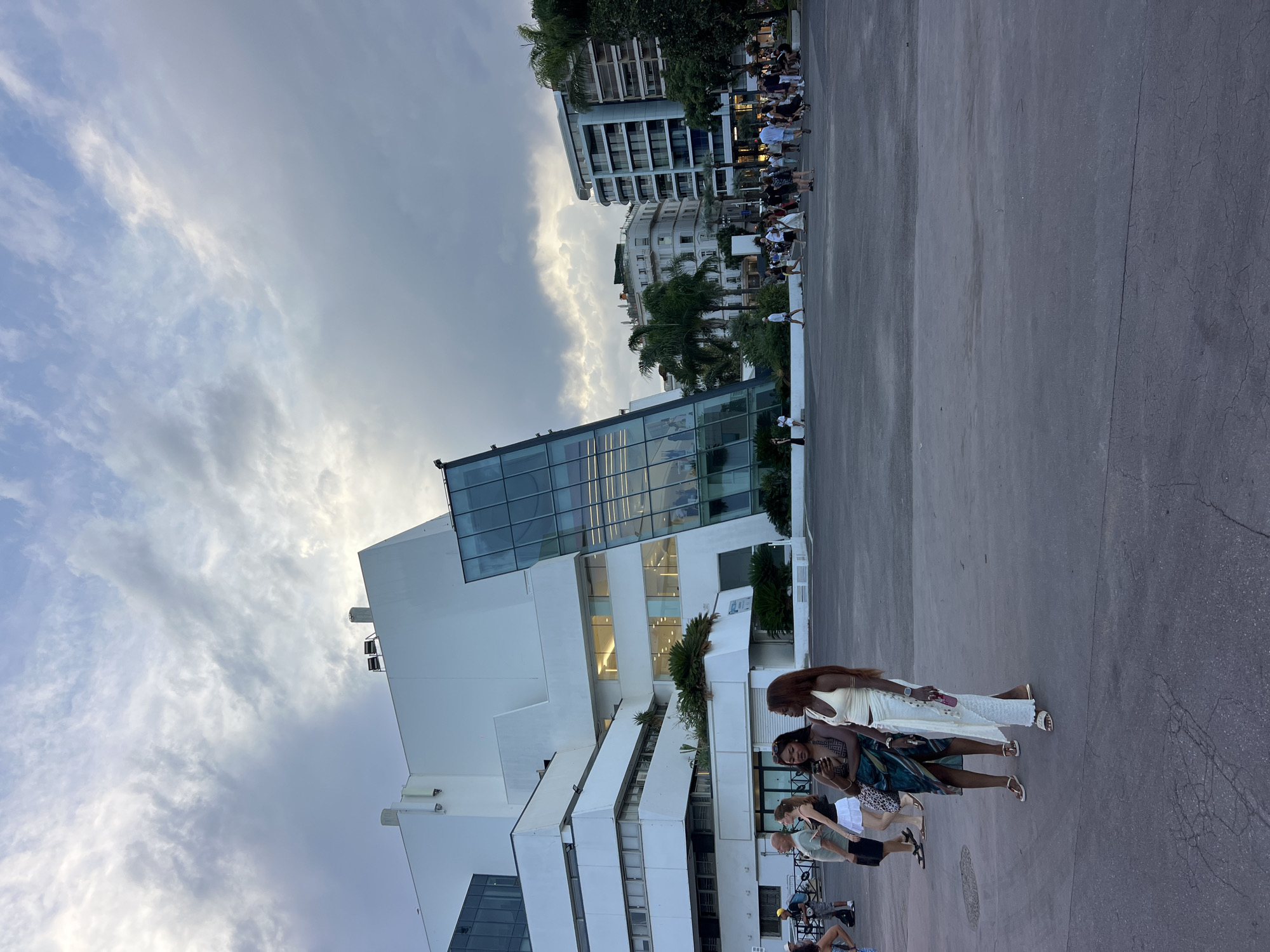 The image shows the Palais des Festivals et des Congrès in Cannes, France. Two women are standing in the foreground, with a crowd of people in the background. The Palais des Festivals is a large, modern building with a white facade and a glass facade. The sky is cloudy, and the sun is setting. The area in front of the Palais des Festivals is a large, open plaza. The overall scene suggests a public event or gathering at the convention center.
