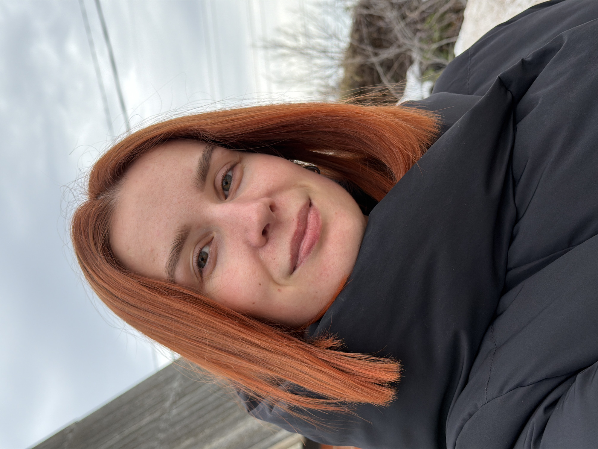 A young woman with red hair smiles at the camera. She is wearing a black puffer jacket. The background includes a cloudy sky, trees, and a snowy area. A grey roof can be seen at the bottom left of the image. The location is likely outdoors in a cold climate. No specific details confirm it is in Chaikovsky, Russia.