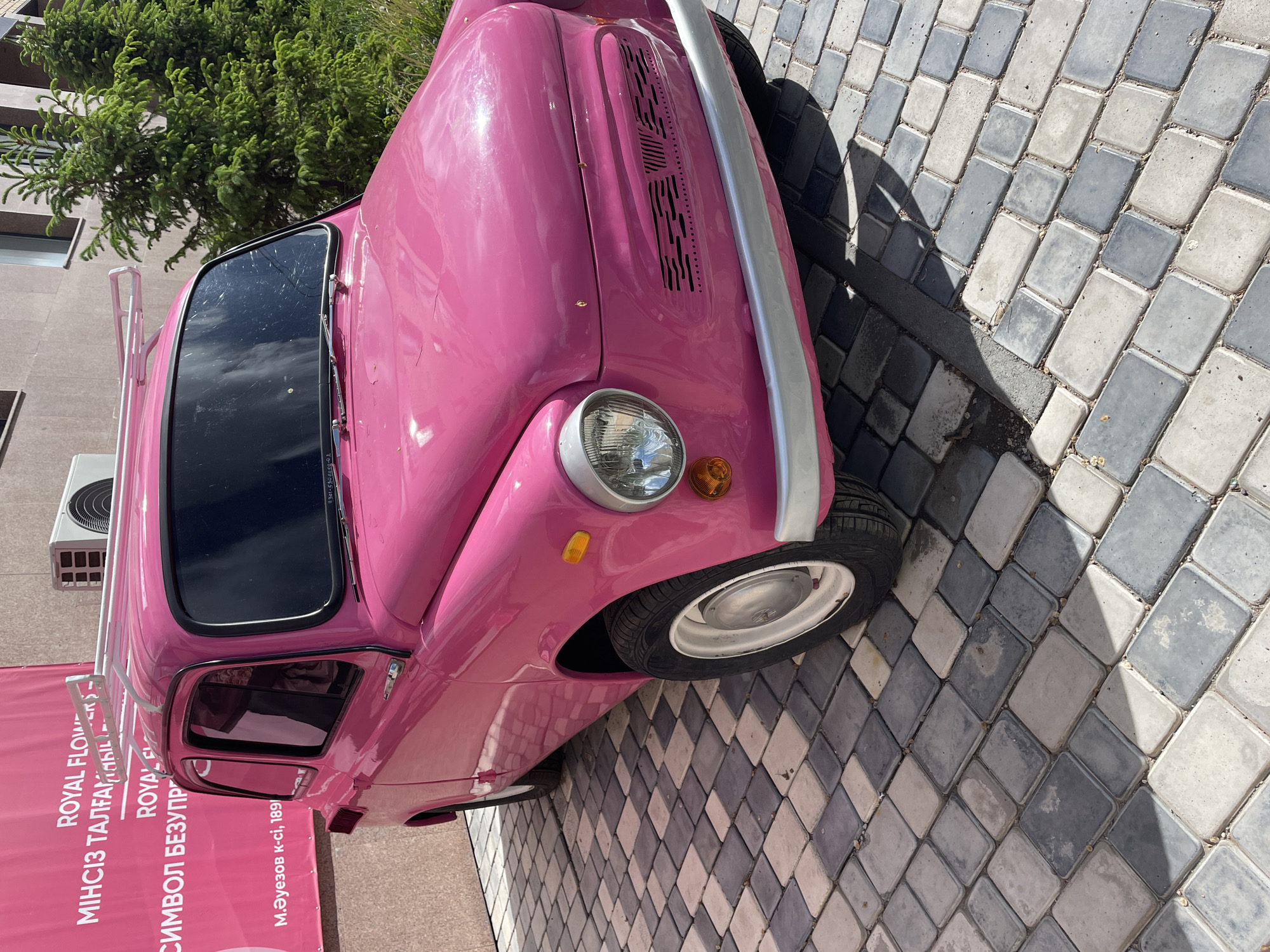 Here is a description of the image:
A bright pink vintage car is parked on a patterned brick surface. The car is facing toward the top left of the frame. It has a silver bumper, white-rimmed tires, and a round headlight with a small orange turn signal light beneath it. The car is parked next to a pink banner with white lettering, which appears to be a sign for “ROYAL FLOWER” in both a Cyrillic language (Kazakh or Russian) and English. The address “М.Әуезов к-сі, 189” is visible on the banner. An air conditioning unit is visible on a wall behind the car. A tree with dense green foliage is partially visible behind the car. The patterned brick surface shows an array of gray and white hues. Based on the address visible on the banner, the location is M. Auezov Street, 189, in Kokshetau, Kazakhstan.