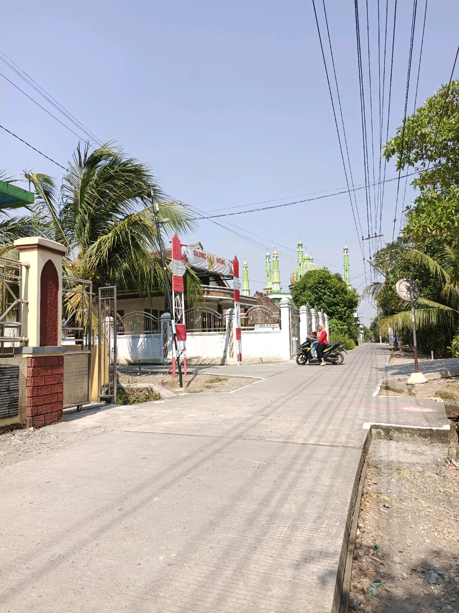 This is a photograph of a street scene in Cangkring Rembang, Indonesia, on a bright, clear day. The street is paved with concrete and appears to be a residential or semi-commercial area. To the left, there is a building with a red and white striped decorative element and a metal gate. Behind the gate, a palm tree is visible. Further down the street on the left, there is a white building with a metal fence.
Dominating the background is a mosque with several green domes and minarets. In front of the mosque, there is a white fence. On the right side of the street, there are trees and utility poles with numerous overhead wires.
In the foreground, a person wearing a red top and dark pants is riding a motorcycle with another person, also wearing dark pants, as a passenger. They are traveling away from the camera, down the street. The motorcycle is black and silver. The road surface shows tire marks and some debris. The overall impression is a typical street view in a sunny, tropical environment.