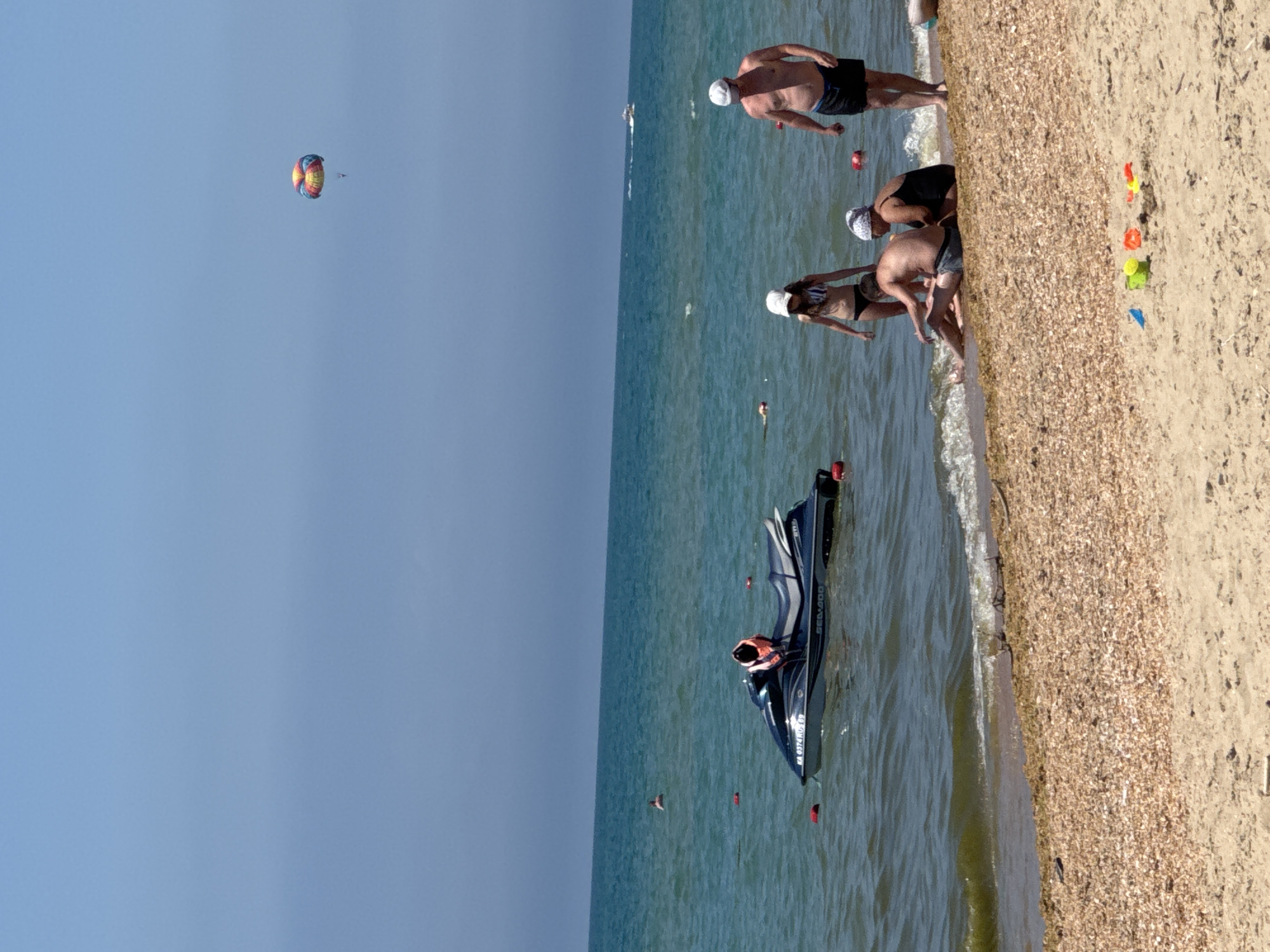 The image depicts a beach scene in Vityazevo, Russia. The foreground is a sandy beach with small pebbles and seashells. In the middle ground, several people are visible near the water's edge. Two men and two women are either sitting or standing. One woman is wearing a black swimsuit and a headscarf, and the other is in a striped bikini and a white hat. One man is wearing black swim trunks, and another is in grey swim trunks. In the water, a dark blue jet ski with an orange life vest on it is floating. Further out in the sea, a person is visible swimming. In the background, the blue sea stretches to the horizon under a clear blue sky. A colorful parachute is visible in the sky, suggesting a parasailing activity. A small boat is also visible in the distance on the water. Scattered on the sand are a few children's toys.