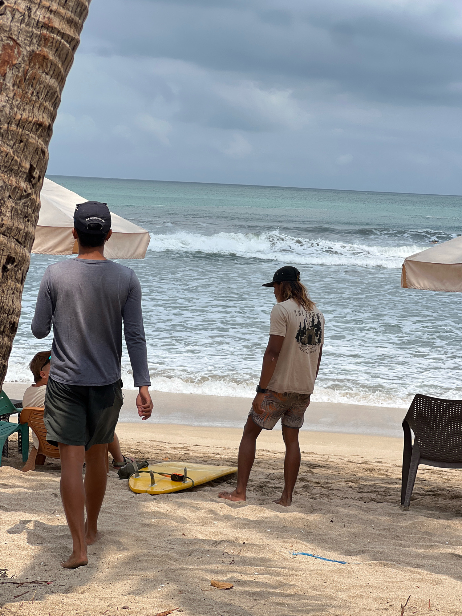 A wide shot captures a beach scene in Kuta, Indonesia, under a cloudy sky. In the foreground, a palm tree trunk partially obscures the left side of the frame. Two young men stand on the sand, facing away from the camera towards the ocean. The man on the left wears a grey long-sleeved shirt and dark shorts. The man on the right is dressed in a light-colored t-shirt with a graphic design and patterned shorts. A yellow surfboard rests on the sand between them. Further back, a person is seated on a chair under a beach umbrella. The ocean has moderate waves, and the beach is composed of light-colored sand. A dark chair sits on the right side of the image.