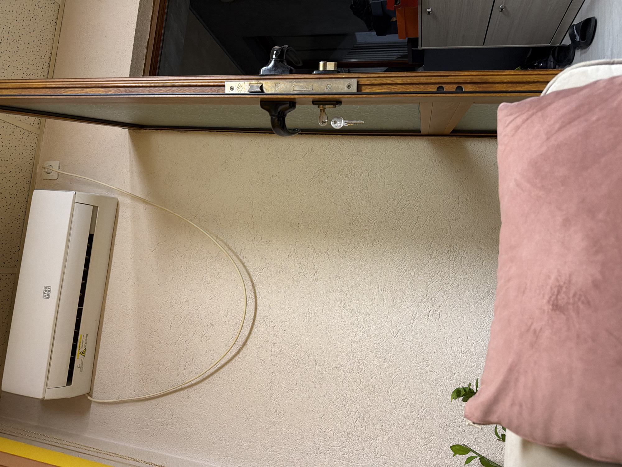 A close-up, indoor shot shows a wall with an air conditioning unit. The unit is white and mounted high on the wall. A power cord runs from the unit and curves along the wall. Above the wall is a shelf with a wooden frame. A door is partially visible above the shelf. The door has a latch and key in the lock. In the foreground, a pink pillow rests on a white surface. Green leaves from a plant are visible near the bottom right corner. The location appears to be a room inside a building. Panagyurishte, Bulgaria is not discernible from the given information.
