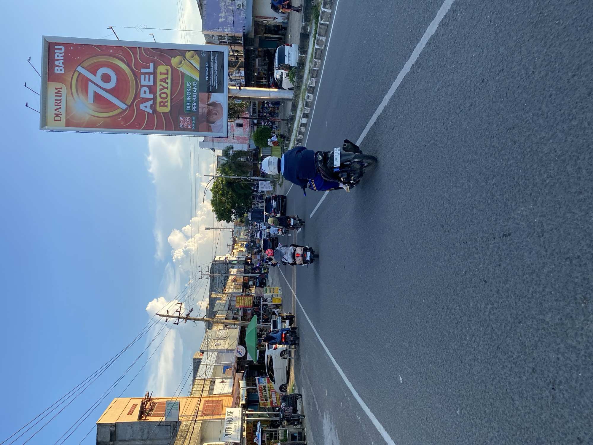 A wide, asphalt road in Lhokseumawe, Indonesia, is shown from the perspective of a person on a motorcycle. The road is lined with buildings on both sides, some appearing to be shops and others residential. A large billboard advertising "Djarum Baru 76 Apel Royal" is prominently displayed on the right side of the road. Several people are seen riding motorcycles, moving in the same direction as the viewer. The sky is blue with scattered clouds, and the scene appears to be during daylight hours, likely late afternoon given the lighting. The environment depicts a typical urban street scene with traffic and commercial establishments.