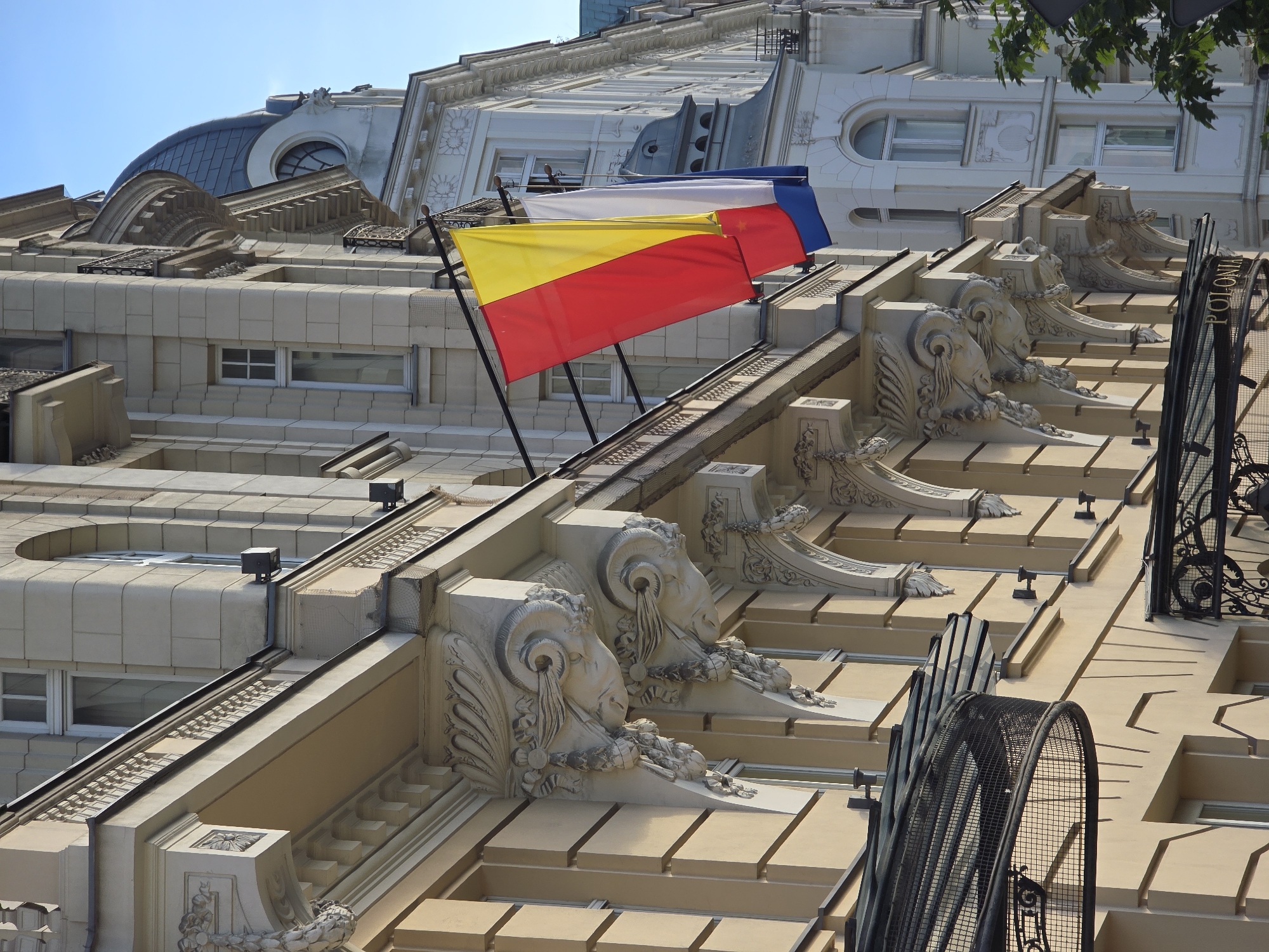 The image shows an upward view of ornate building facades in Warsaw, Poland, under a clear blue sky. The architecture is characterized by cream-colored stone, intricate decorative elements, and multiple windows. Prominently displayed are three flags mounted on poles: a red and yellow striped flag below a white flag, and a blue flag above the white one. These appear to be the flags of Romania (red, yellow, blue), though they are arranged horizontally.
The building's exterior features decorative brackets shaped like ram heads, ornate stonework around windows, and carved floral motifs. Several windows with white frames are visible, some with multiple panes. Metal railings and fences, including a decorative arched one with the word "POLONIA" visible on it, add to the architectural detail. The scene suggests a bright, sunny day. The focus is on the rich architectural details of the building and the national flags, creating a sense of cultural representation and historical grandeur.