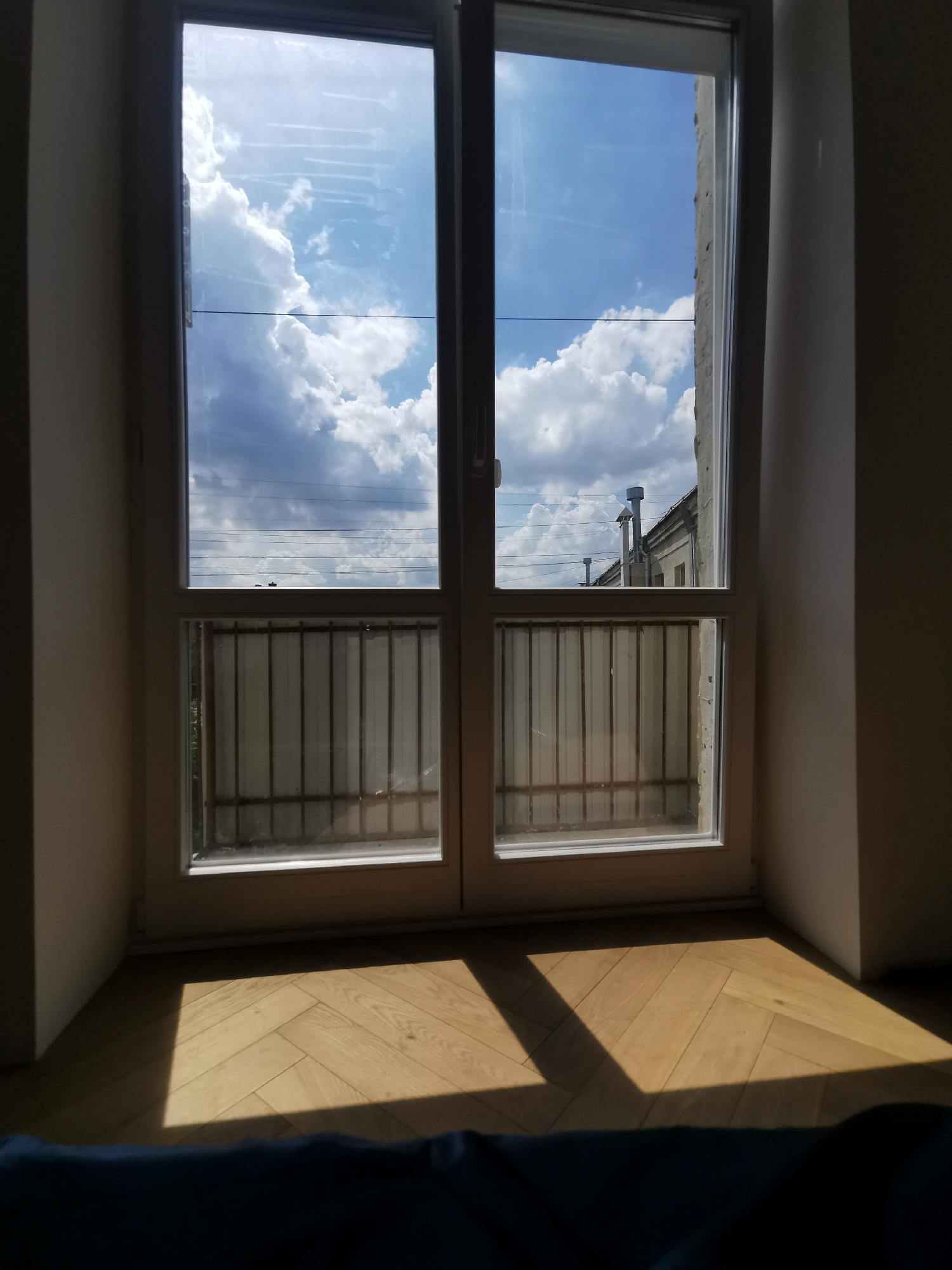 A sunlit interior view shows a double-paned window, revealing a cloudy blue sky and an exterior view of a building with chimneys. Below the window's upper panes are lower sections with metal bars. Sunlight casts a shadow on a light-colored herringbone wood floor. The scene is shot from a low angle. No people are visible. The location in the city of Moscow, Russia is not discernable from the image.