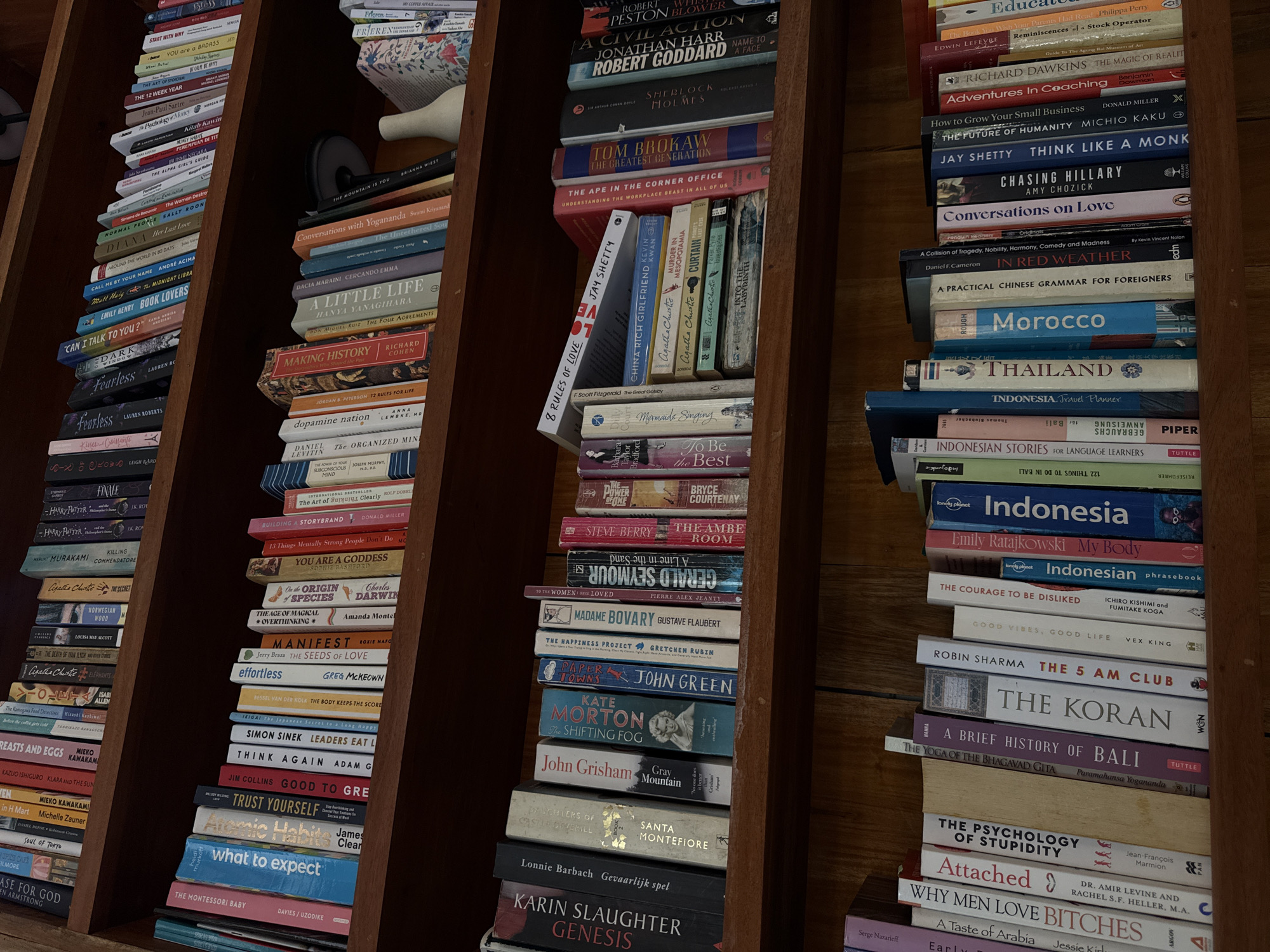 The image shows a collection of books on multiple wooden bookshelves, presumably located in the Ubud District, Indonesia, based on some of the book titles related to Indonesian culture and travel. The books vary in size and subject matter, ranging from travel guides and language learning resources to literature, self-help, and philosophy. The spines of the books are visible, revealing the titles and authors. The scene is indoors, with natural lighting present. There are no people visible.