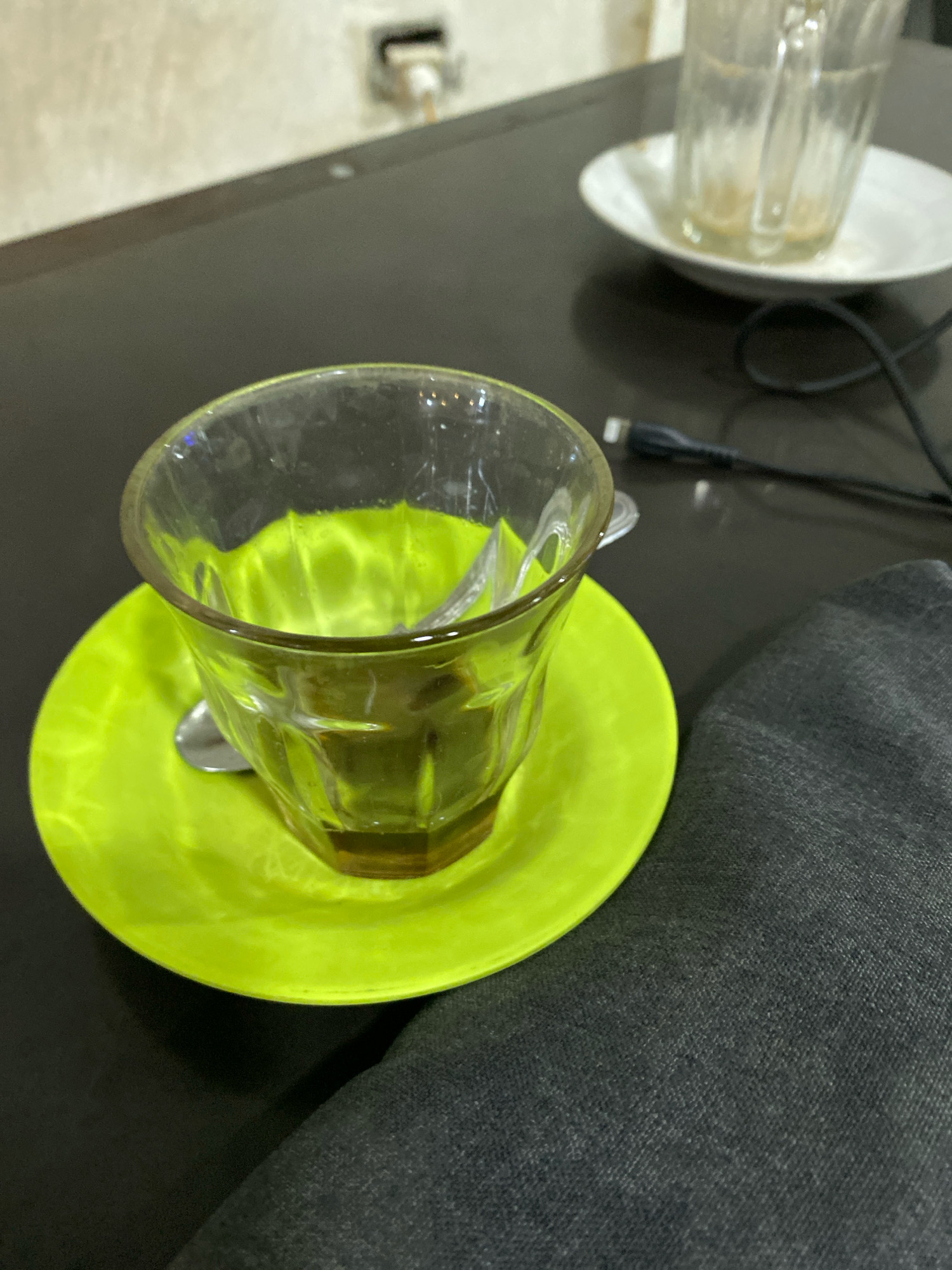 An overhead shot shows a close-up of a green saucer with a clear glass cup filled with a dark beverage. A silver spoon rests inside the cup, and another is visible on the saucer. The cup and saucer sit on a dark, reflective surface that appears to be a table. In the background, another glass and saucer are blurred, along with a black charging cable. The scene is lit from above, creating reflections on the table and the glass. The overall impression is of a coffee break or a moment of pause. No people are visible in the frame. The image does not contain any contextual information that would allow for its placement in Lhokseumawe, Indonesia, or any specific city.