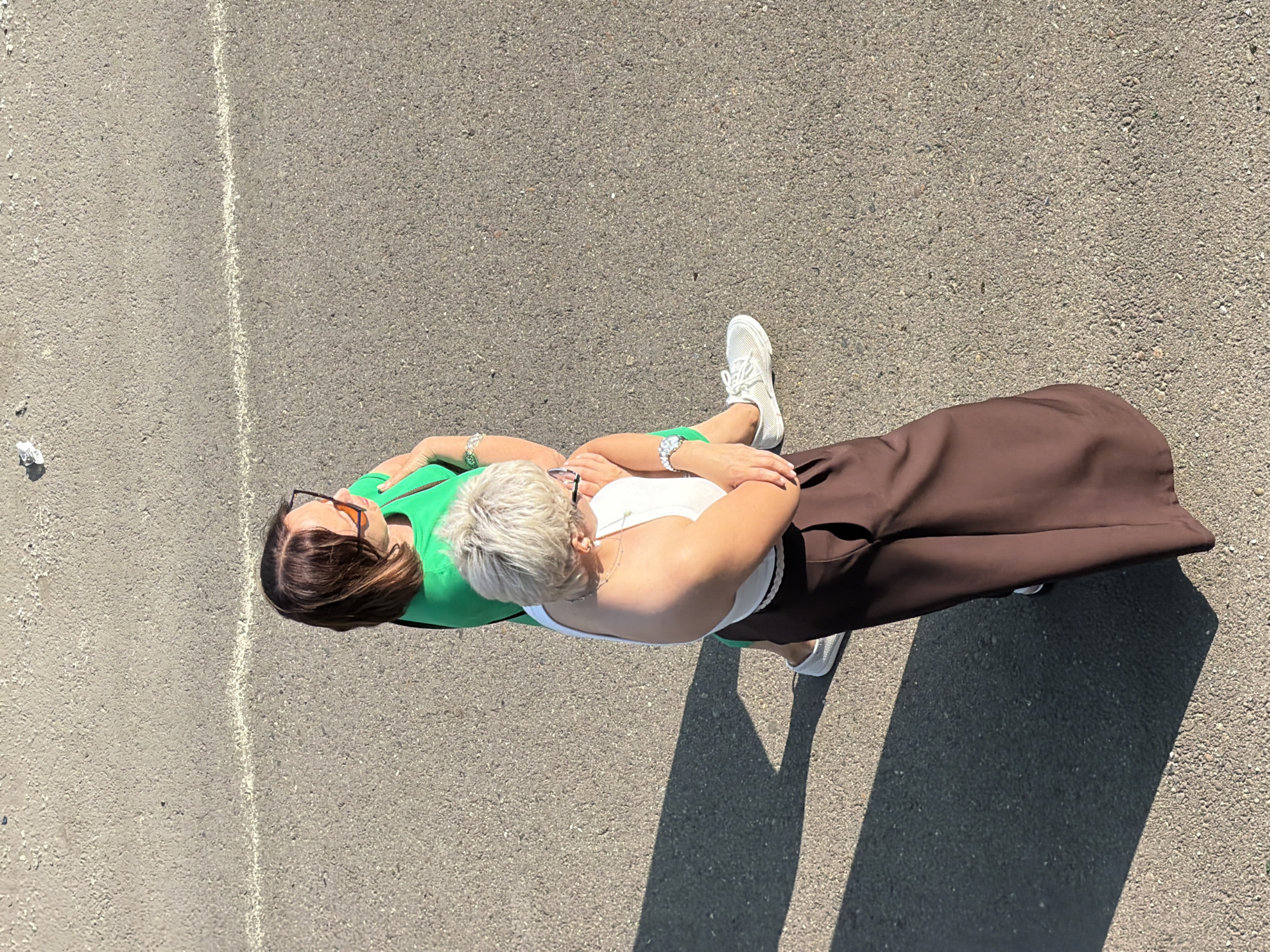 In a high-angle shot taken on an asphalt surface, two women are captured from above. The woman in the foreground, on the right, has short, blonde hair and is wearing a white tank top and wide, brown pants. She has a watch on her left wrist and is holding her arms across her chest. The woman behind her, on the left, has medium-length, brown hair and is wearing a bright green dress and orange-tinted sunglasses. She is also wearing a watch and jewelry on her left wrist. Both women are wearing white sneakers and appear to be standing together. The ground is a textured asphalt, with a faint white line running horizontally across it. A shadow is cast from the left, obscuring part of the asphalt. There is a small piece of white debris visible on the asphalt in the upper left corner of the image. The lighting suggests it is daytime, with the sun casting strong shadows. The scene is likely an outdoor urban setting.