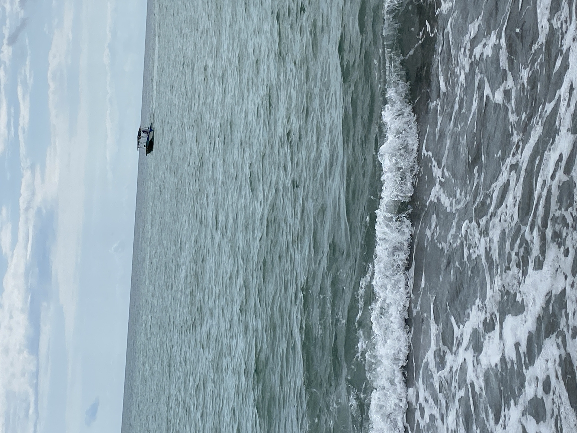A medium-sized boat with a canopy is navigating the choppy, grey sea under an overcast sky. The boat appears to have at least one person on board. Waves are breaking near the shore, creating white foam that washes onto a dark, sandy beach. The horizon is visible in the distance, separating the sea from the sky. The scene is likely located on the coast of Sokhumi, Abkhazia.