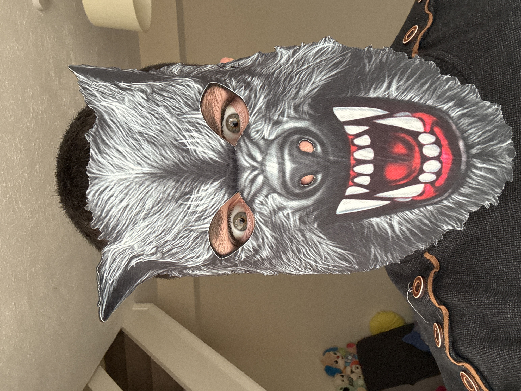 A person is wearing a wolf mask. The person is wearing a dark gray top with copper-colored trim and decorative circles. The background includes a staircase, a wall, and toys. The location is not discernible from the image.