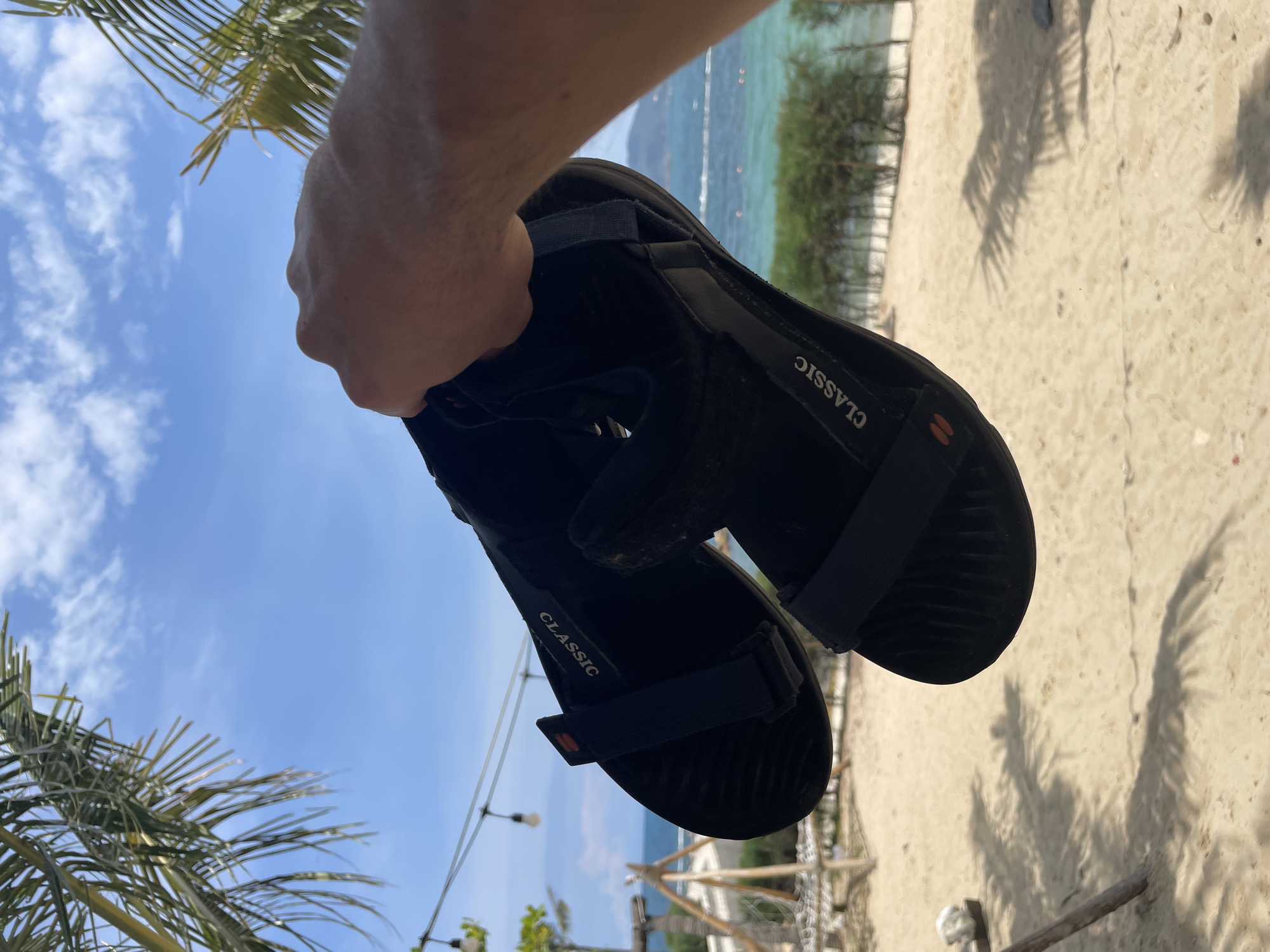 A person is holding a pair of black "Classic" sandals in their hand. The background shows a beach setting with sand, turquoise ocean water, trees, and a partly cloudy sky. The location is suggestive of a tropical beach scene in or near Bac Nha Trang Ward, Vietnam. The angle suggests the photo was taken looking upwards.